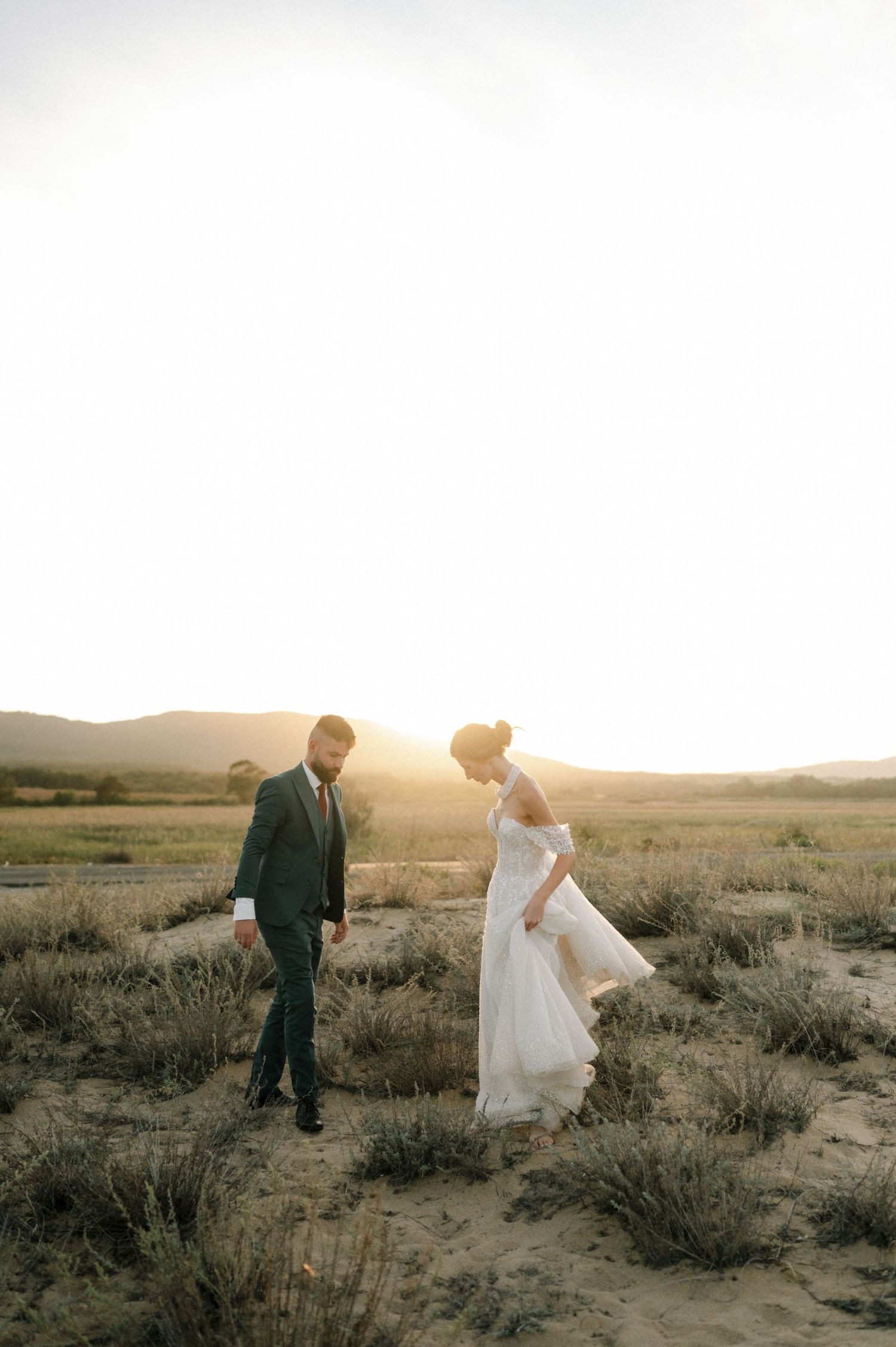 Bride and groom walking through a golden desert field at sunset, bride in flowing white gown, groom in dark suit.