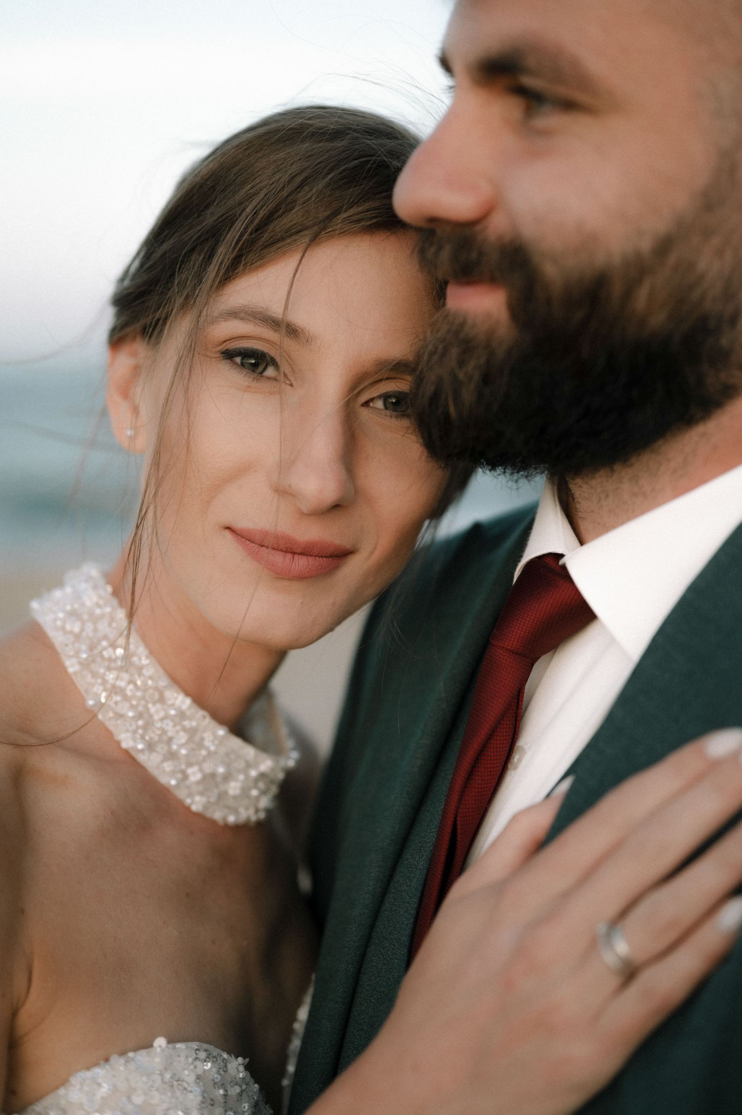 A bride and groom share an intimate moment, she wears a beaded necklace and diamond ring, he wears a dark suit with red tie.