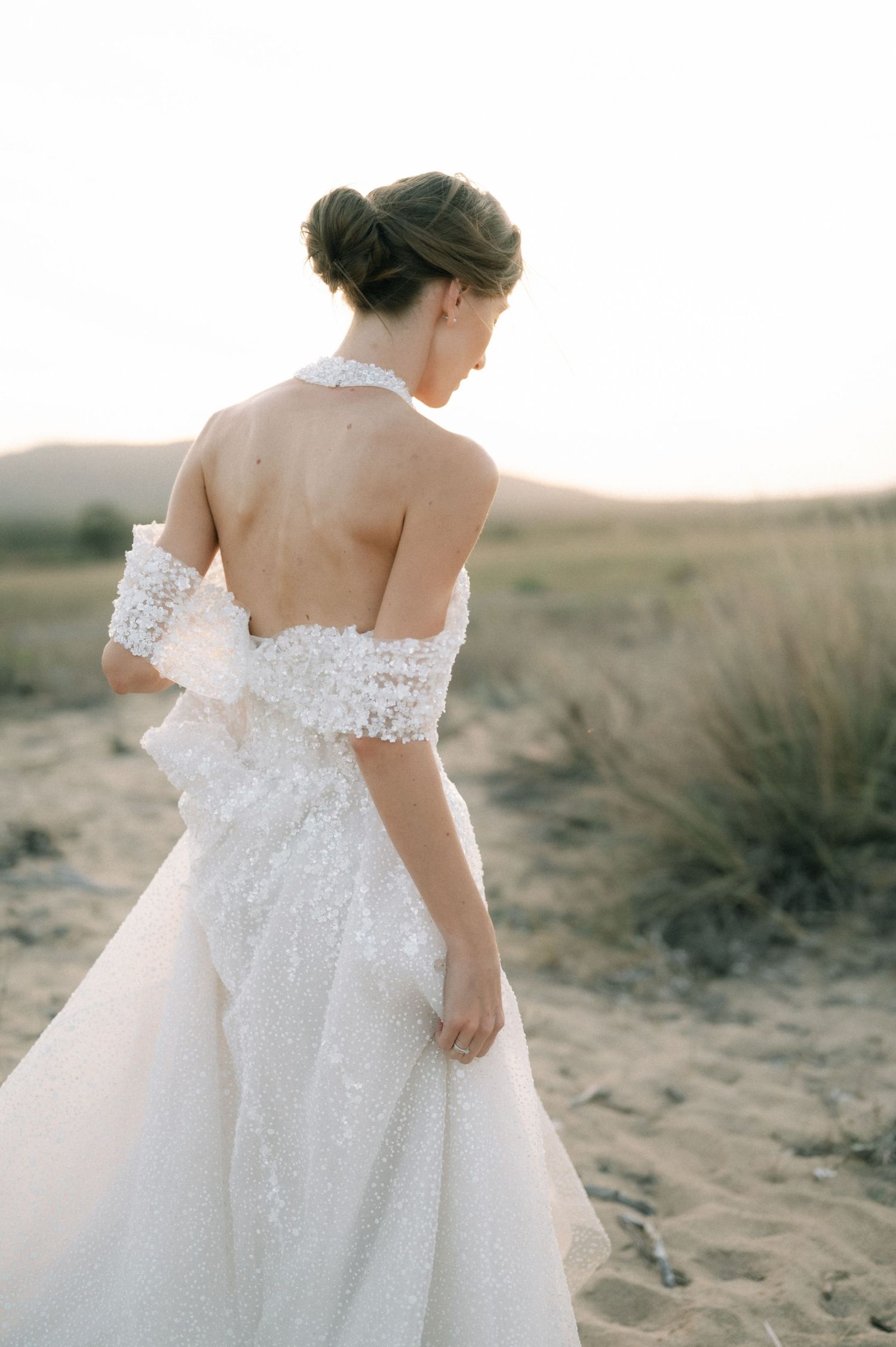 Bride in backless lace halter wedding gown with off-shoulder wrap stands in sandy desert landscape at golden hour.