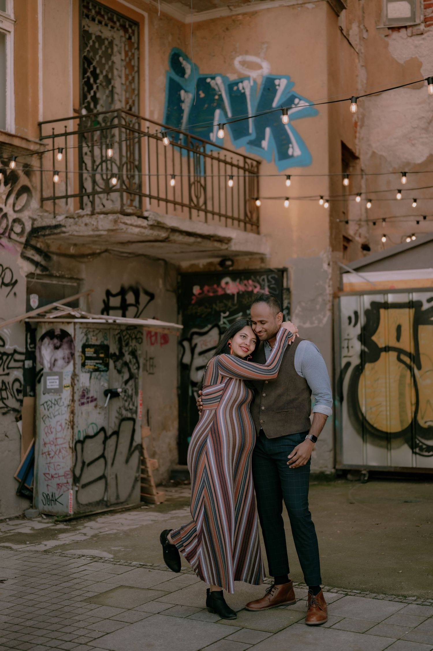 Couple embracing and smiling in an urban alley with graffiti walls and string lights in the background.