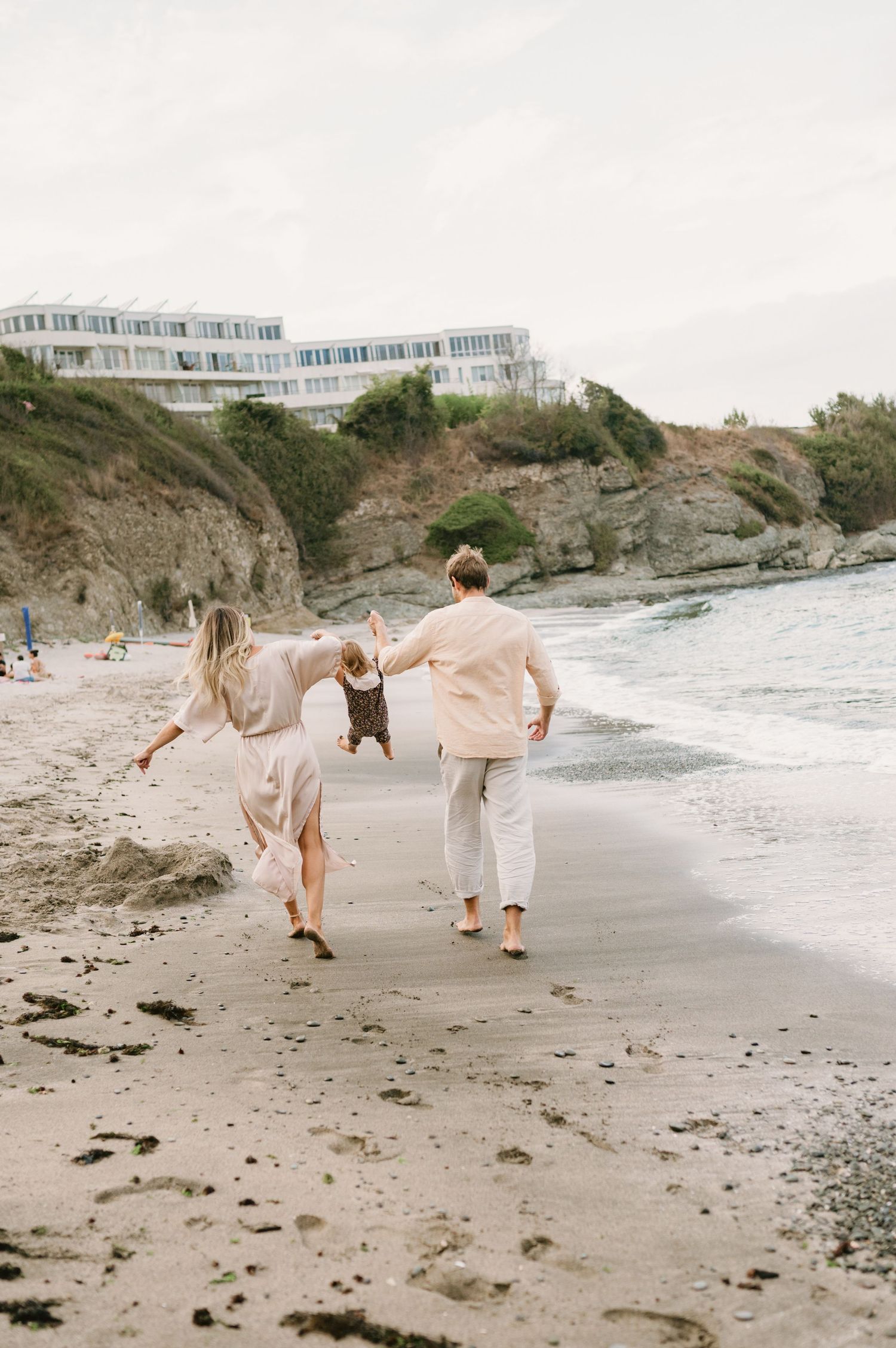 Family walking barefoot on sandy beach near ocean waves, with cliffside hotel in background on overcast day.