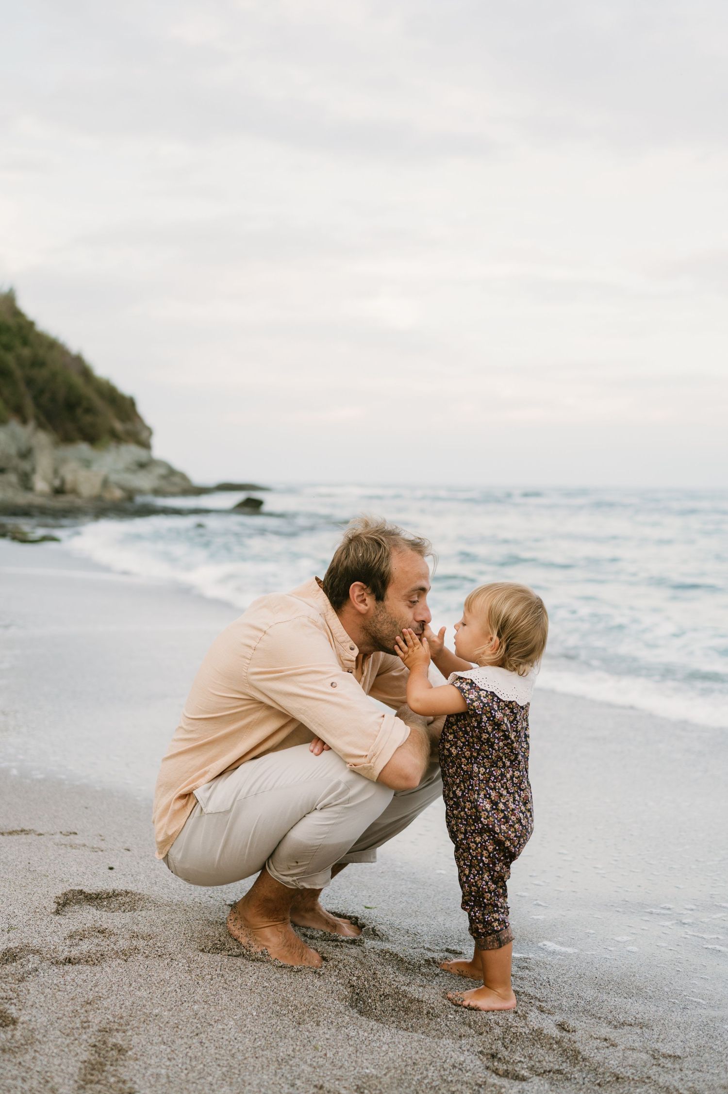 A father crouches on a sandy beach kissing his toddler daughter, with ocean waves and rocky cliffs in the background.