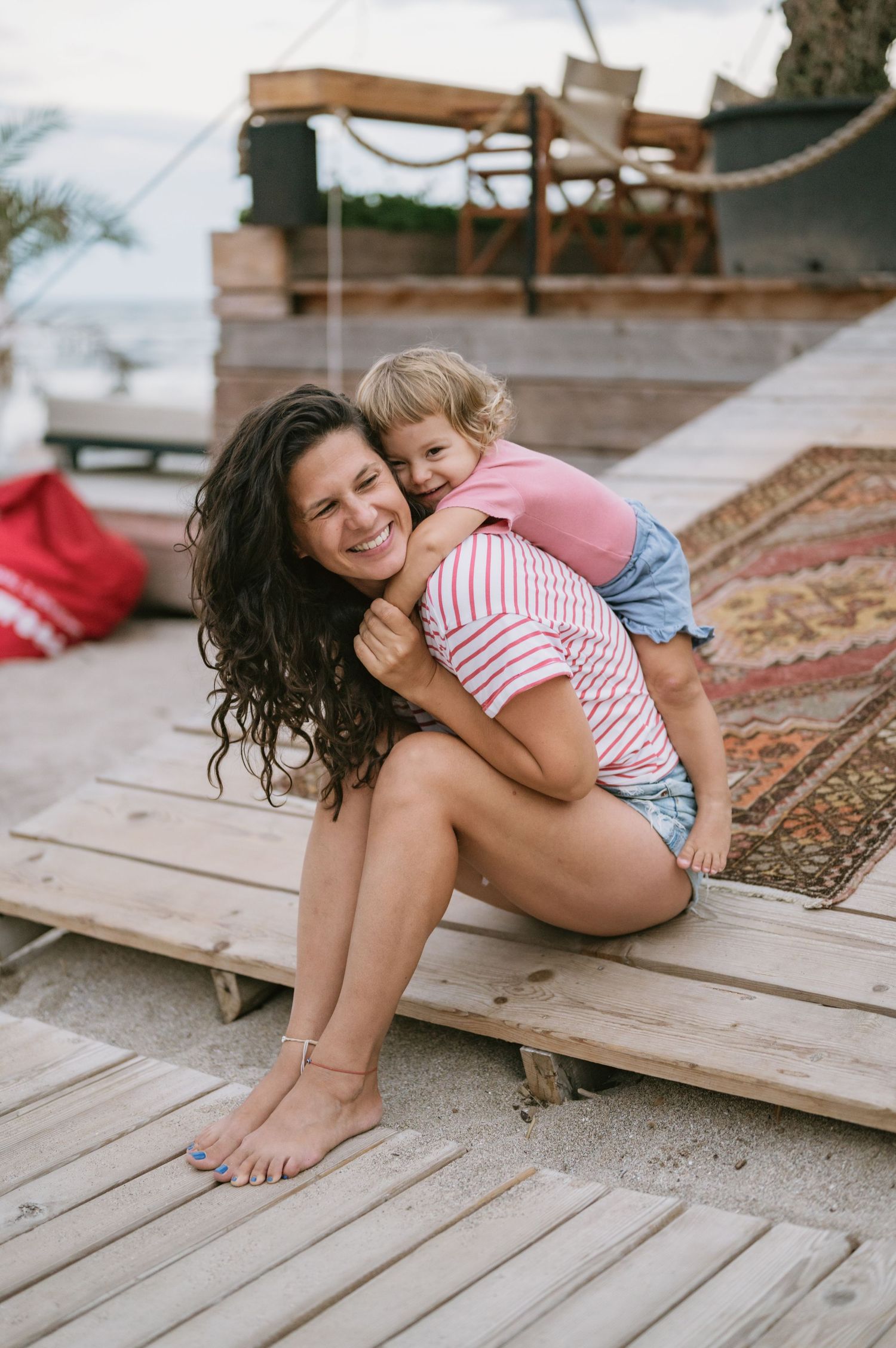 A smiling woman sits on wooden decking at a beach while a happy toddler hugs her from behind.