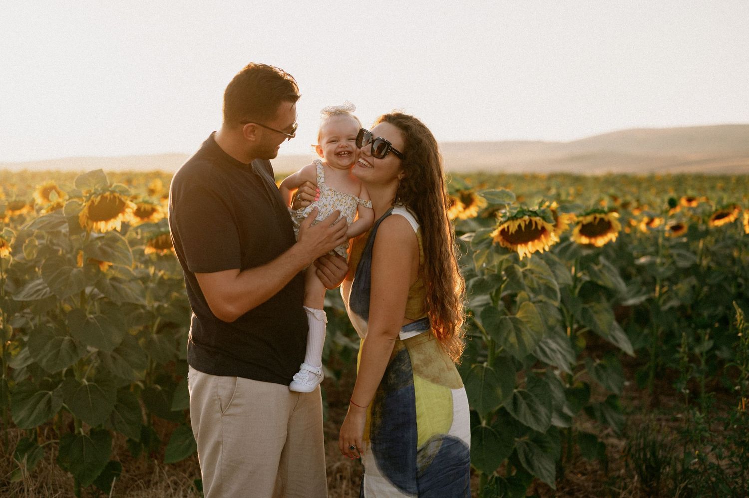 Family with baby posing in sunflower field at golden hour sunset, parents smiling and kissing infant outdoors.