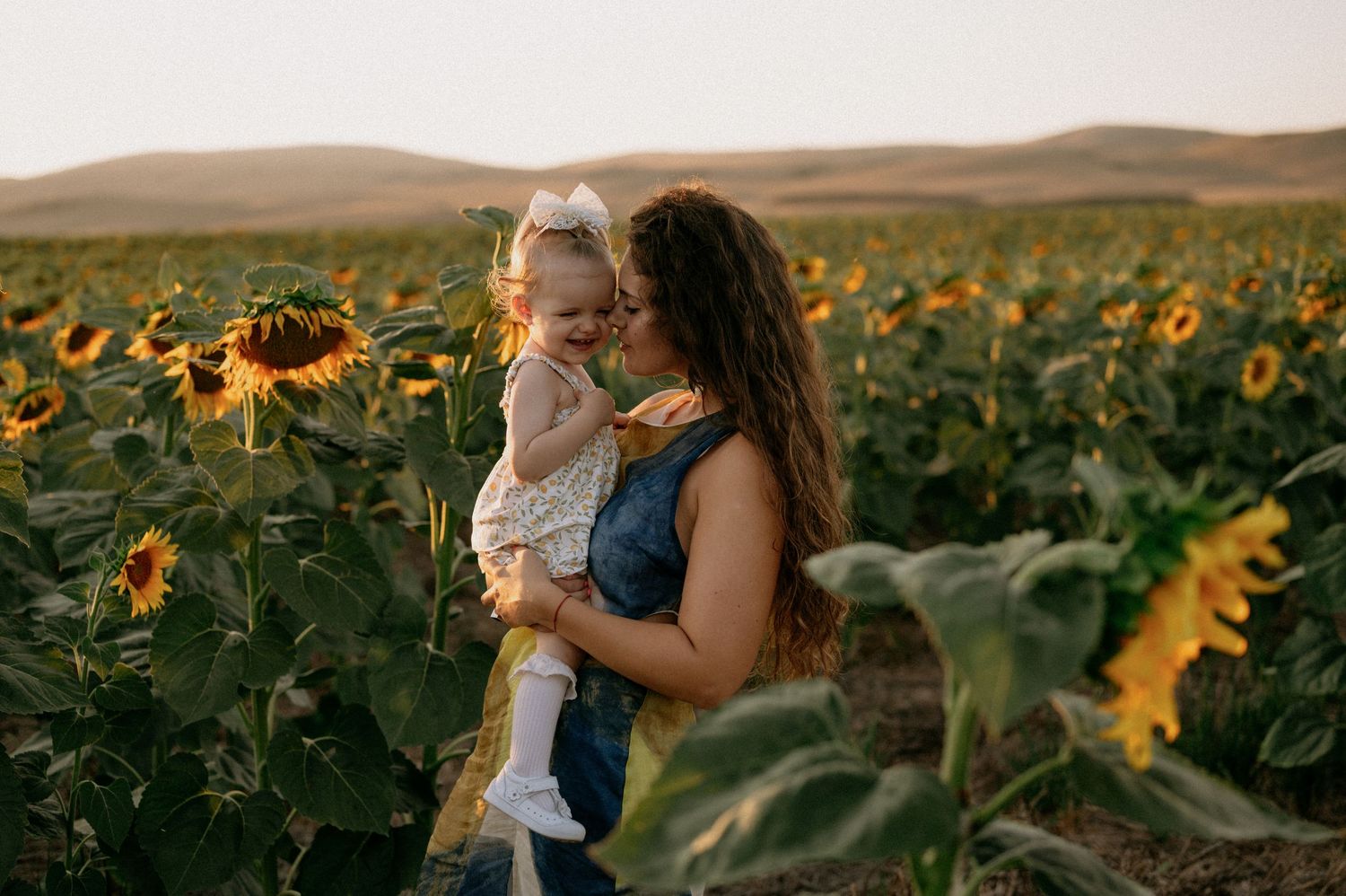 A mother holds her laughing toddler in a vibrant sunflower field at golden hour.