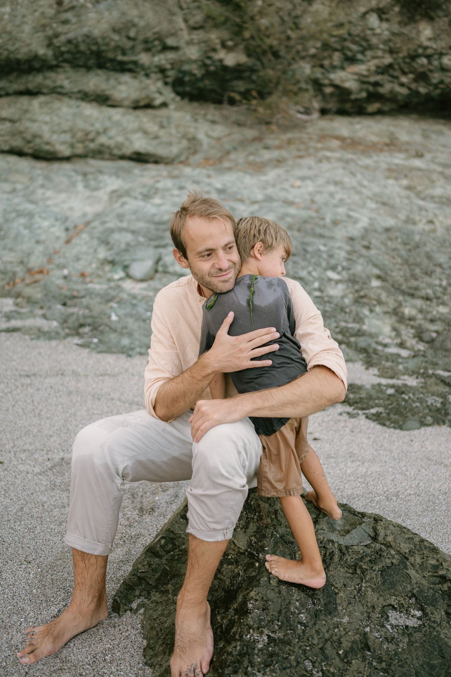 A father sits on a beach rock hugging his young son, with rocky cliffs in the background.