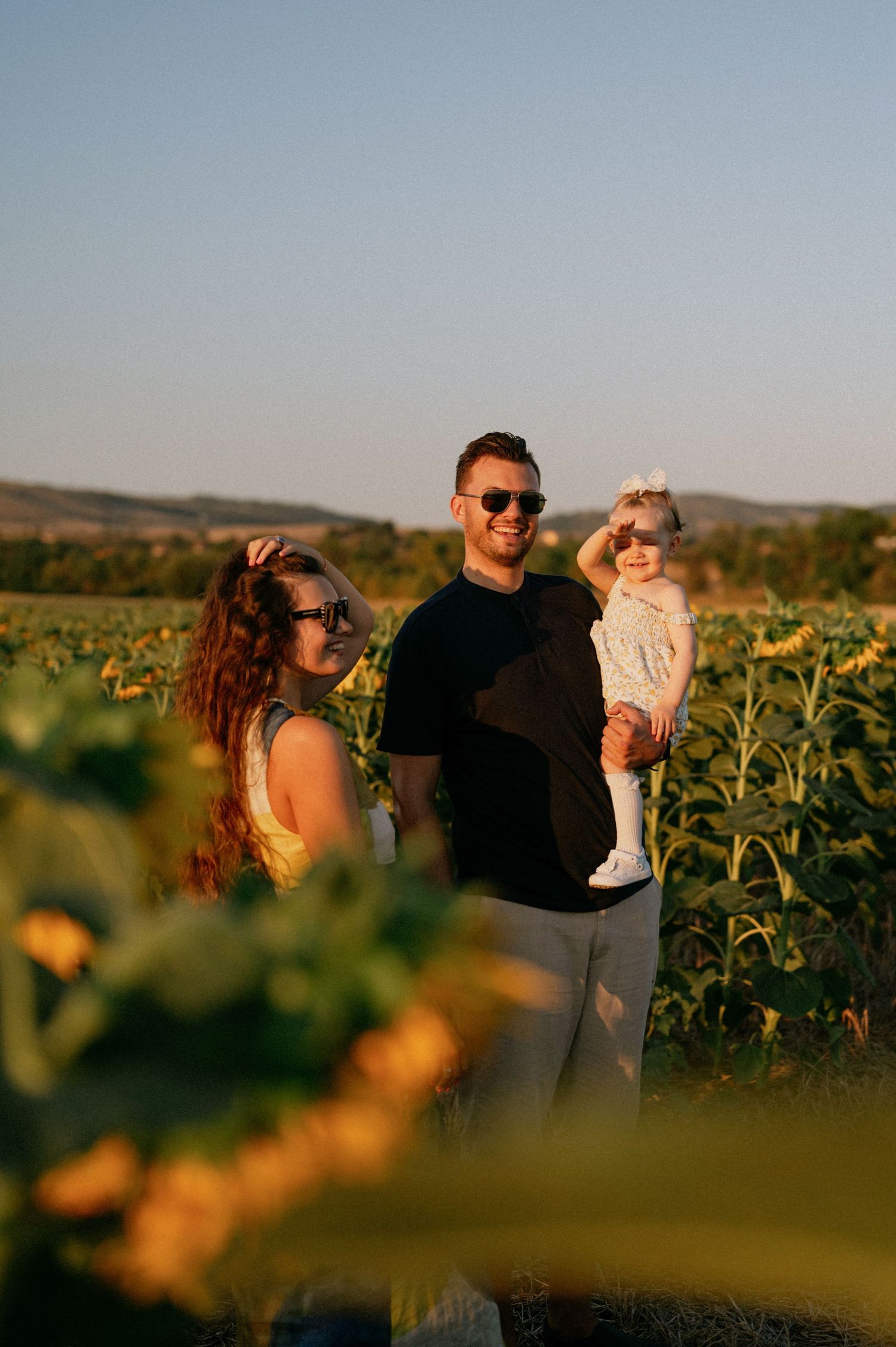 Family of three posing in a sunflower field at golden hour, parents wearing sunglasses, father holding baby girl.