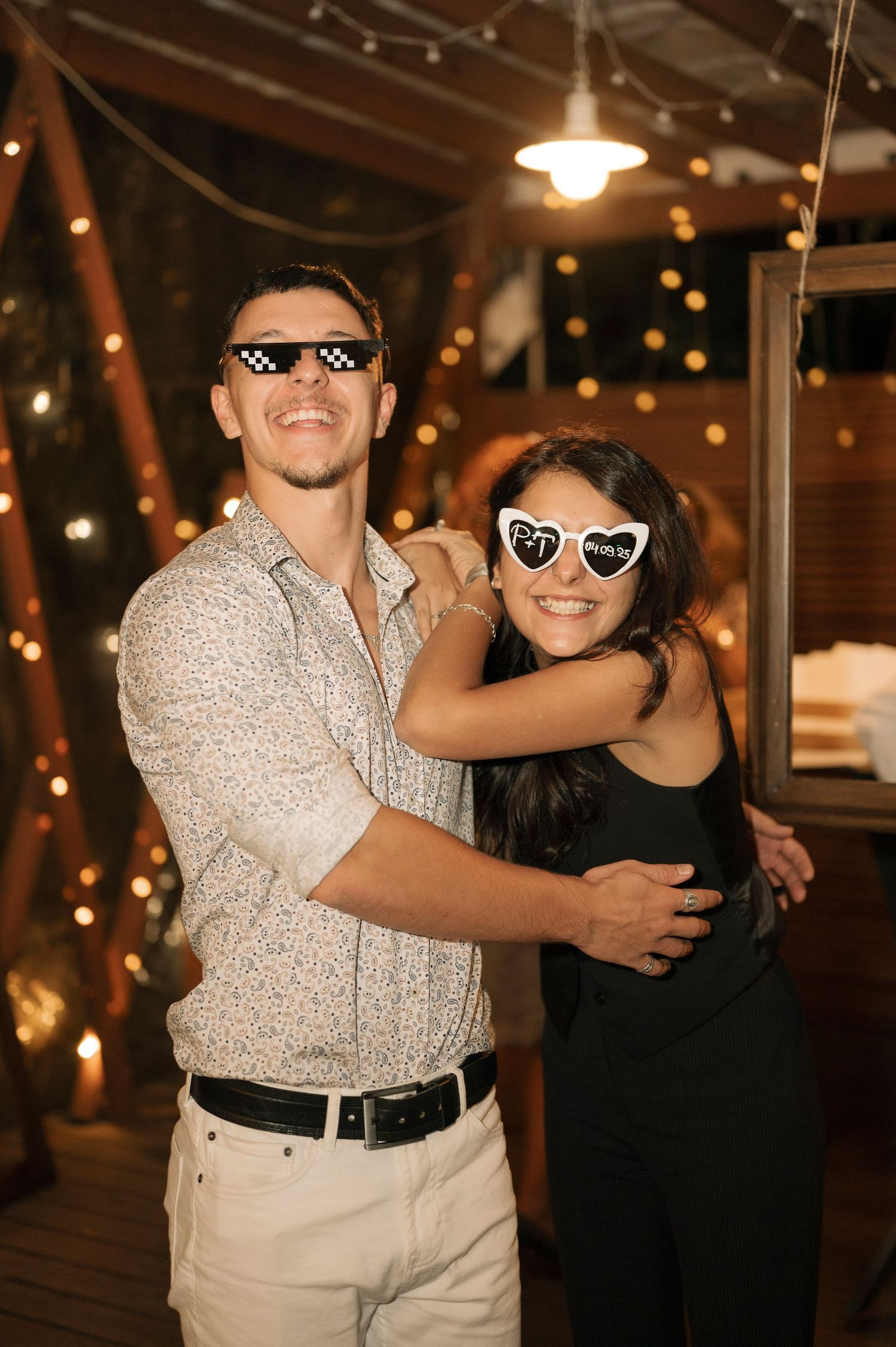 A couple poses together at a party with string lights in the background, showing playful expressions and sunglasses.