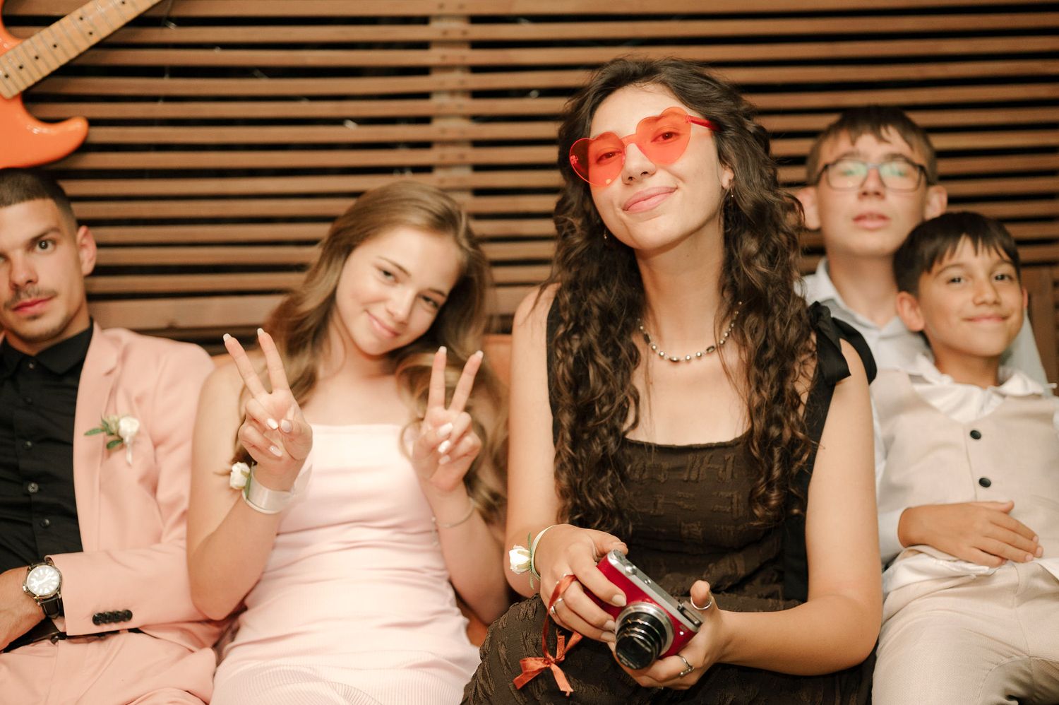 Group of friends at a party with one wearing pink heart-shaped sunglasses and holding a camera in warm lighting.