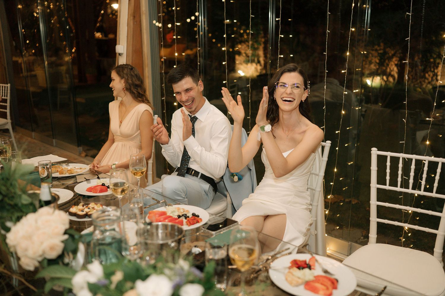A group of wedding guests enjoying dinner at an outdoor reception with string lights overhead and floral centerpieces.