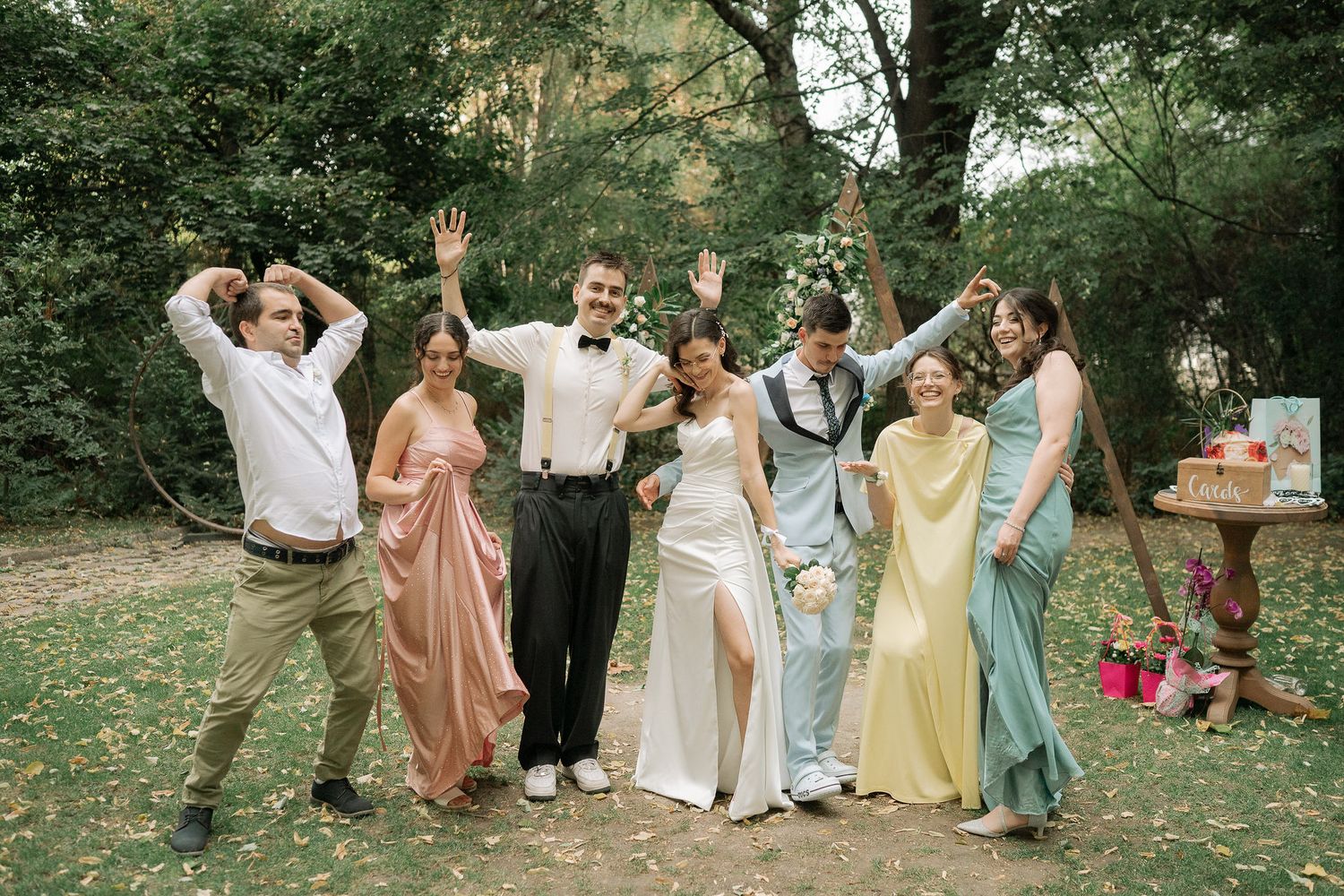 A joyful wedding party celebrates outdoors in pastel formal attire while striking fun poses on a grassy lawn.