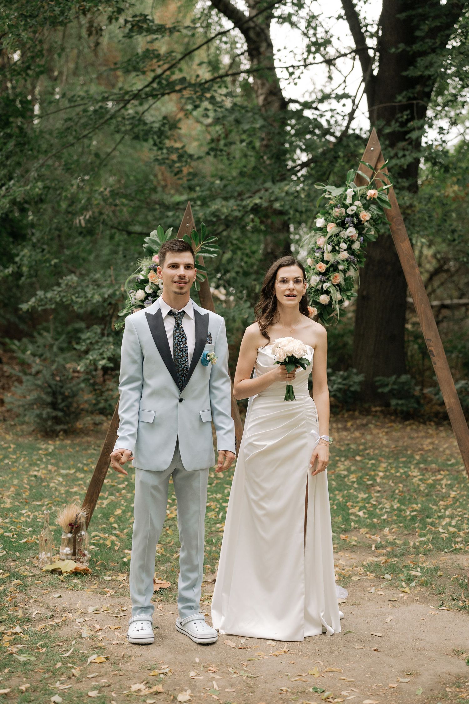 A couple poses for a wedding photo in a wooded outdoor setting with white floral decorations and fall foliage.