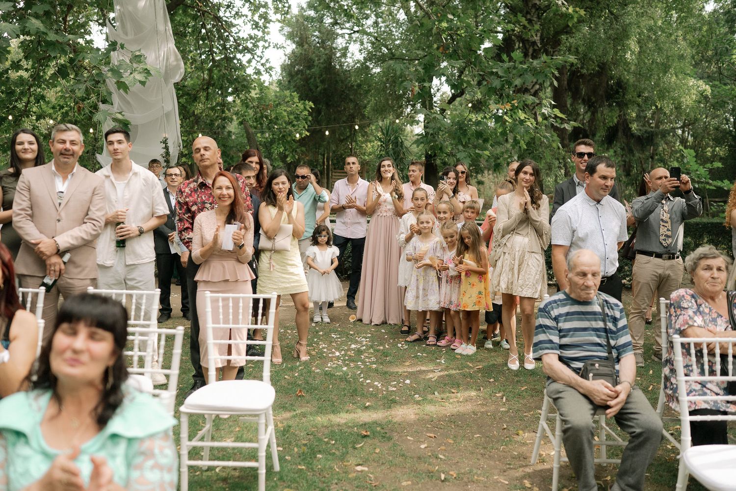 Wedding guests celebrate outdoors in a wooded setting with white chairs and natural decor.