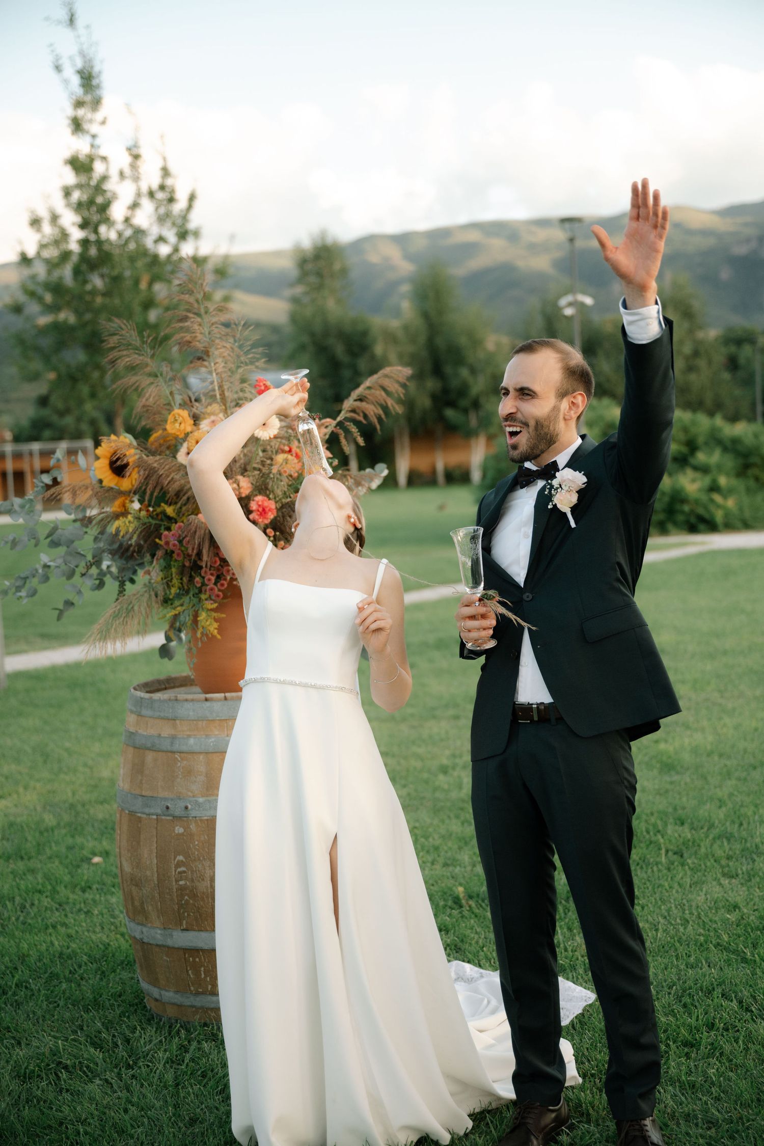 Newlyweds celebrate joyfully on a grassy hillside with wine barrel and sunflower decor against mountain backdrop.