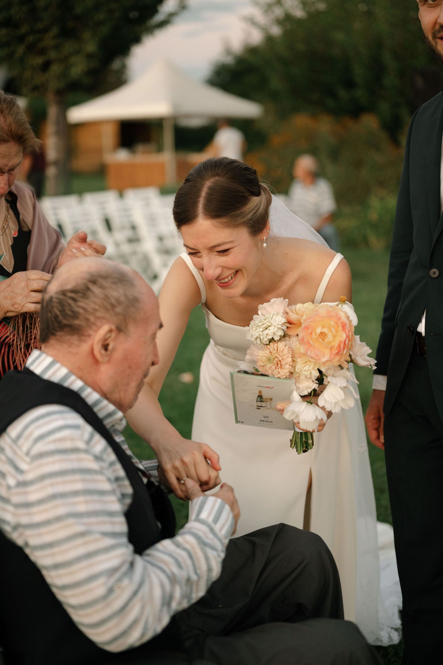 A bride in a white dress holds a peach and cream bouquet while sharing a joyful moment at an outdoor wedding reception.