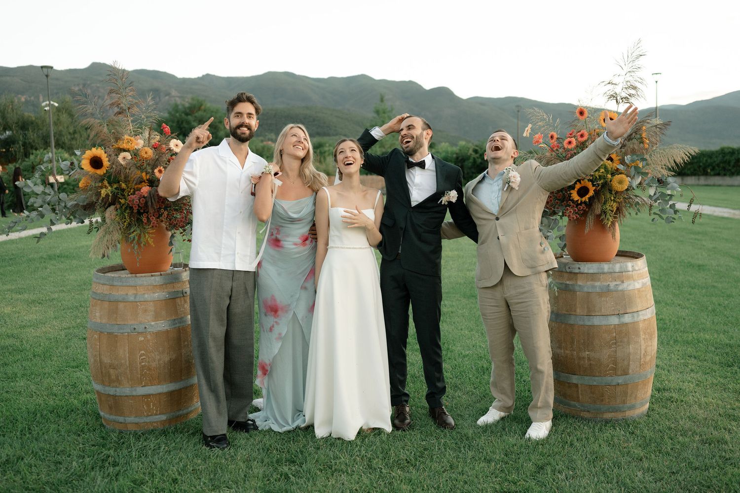 Wedding party poses outdoors at rustic vineyard venue with wine barrels and sunflowers at sunset.