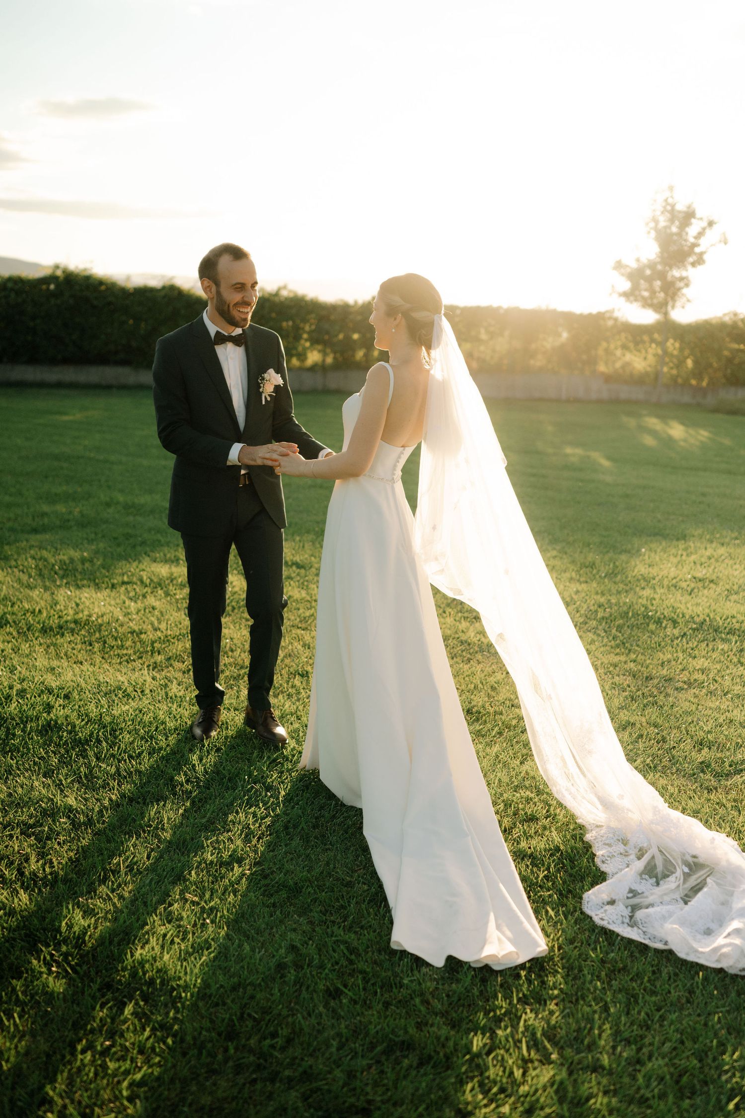 Couple in wedding attire walking through a sunlit meadow during golden hour as the bride's veil trails behind.