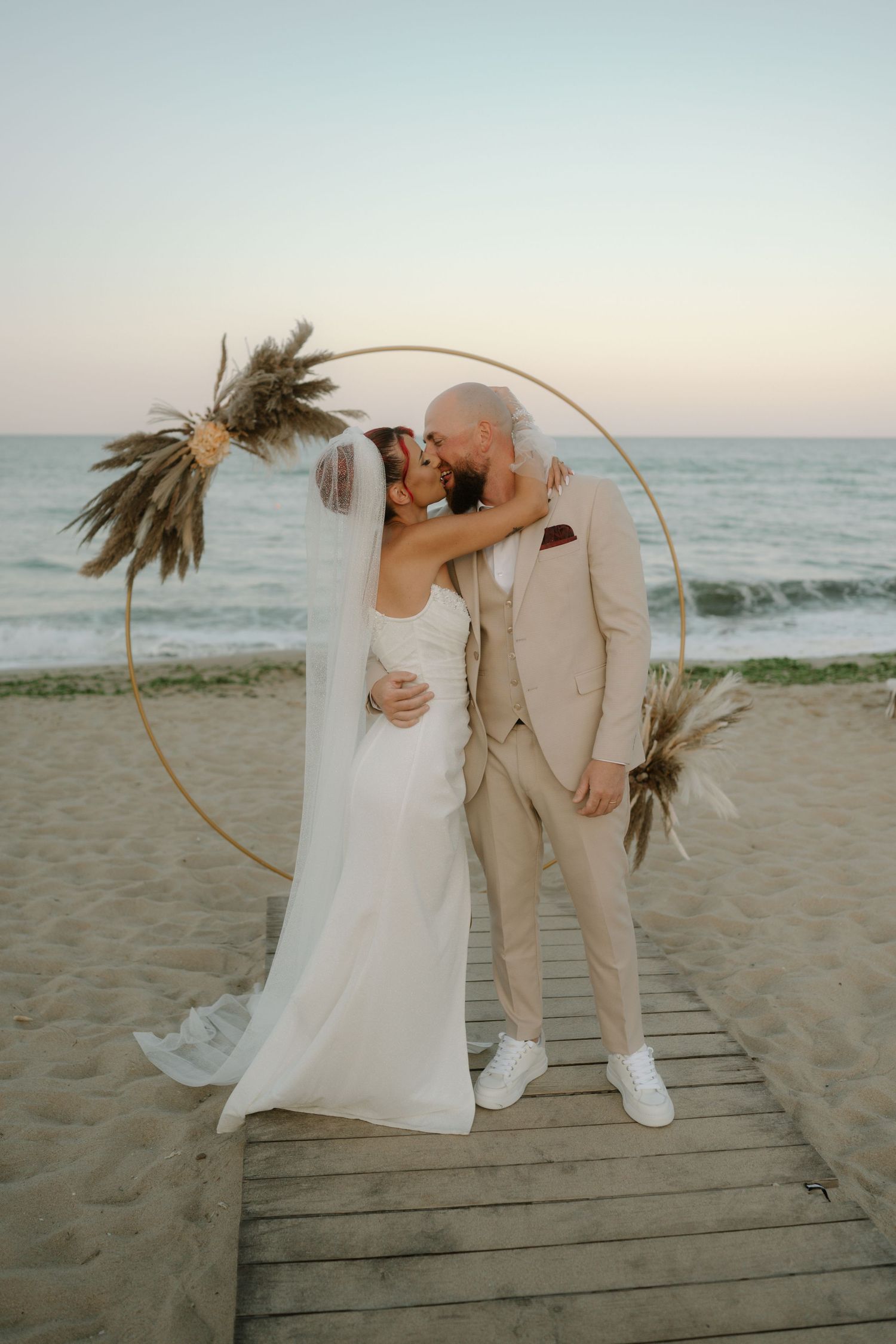 Couple in wedding attire poses on a wooden boardwalk at a beach with palm trees and ocean waves at sunset.
