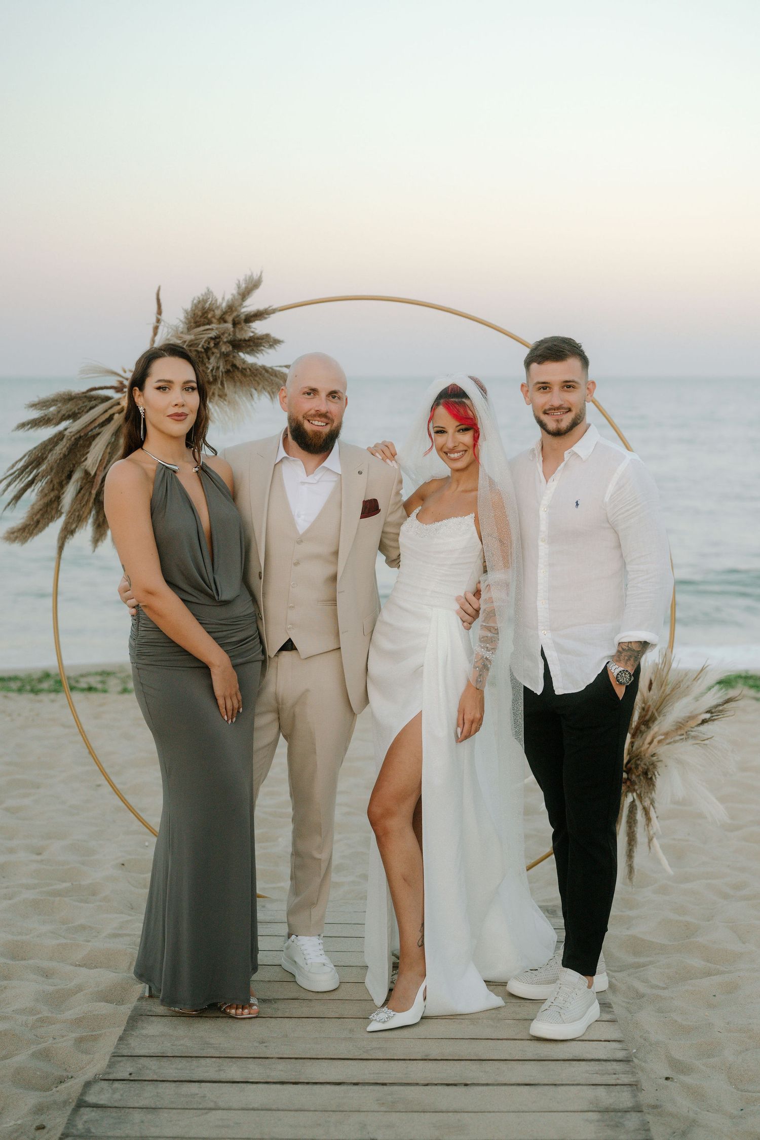 Wedding party poses on wooden boardwalk at beach during sunset with dried palm decor and neutral attire.