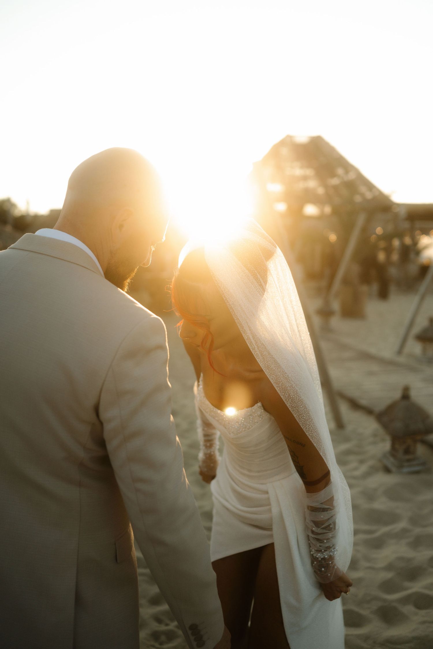 Couple holding hands on a sandy beach at sunset with golden sunlight streaming through their silhouettes.
