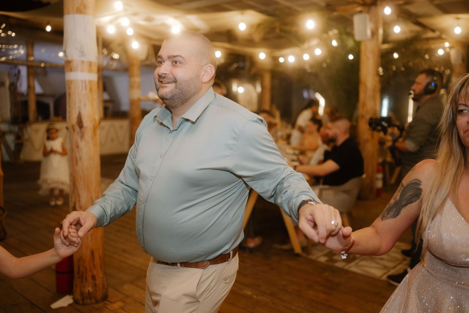 String lights illuminate a rustic indoor venue as wedding guests dance and celebrate during an evening reception.