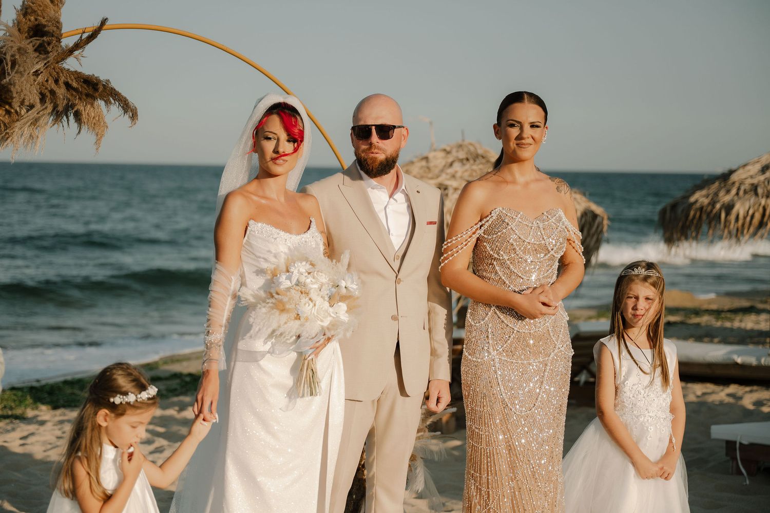 Beachside wedding group photo with light beige and white attire against an ocean backdrop.