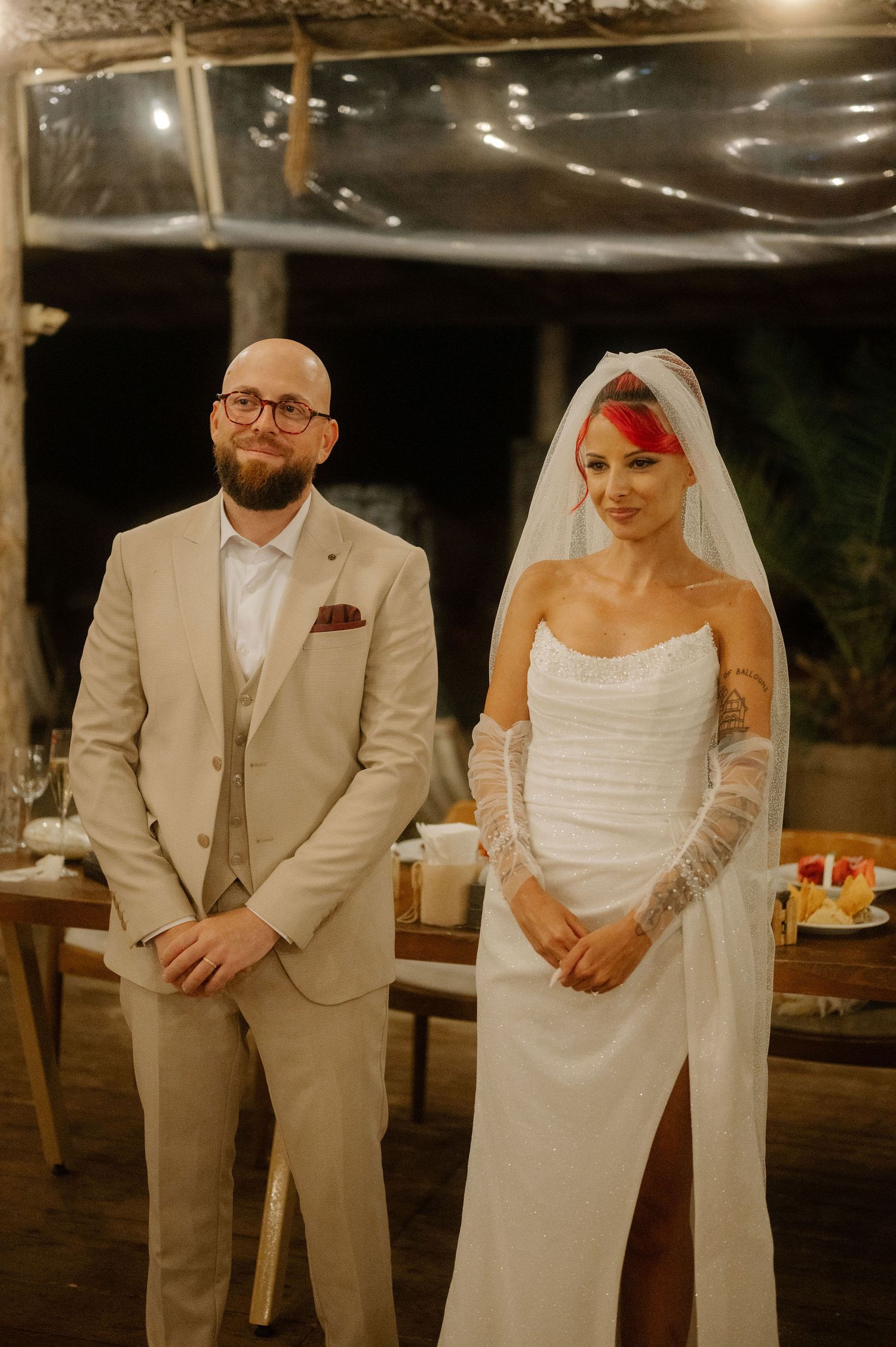 A bride and groom stand together at their evening wedding reception in matching white dress and beige suit.