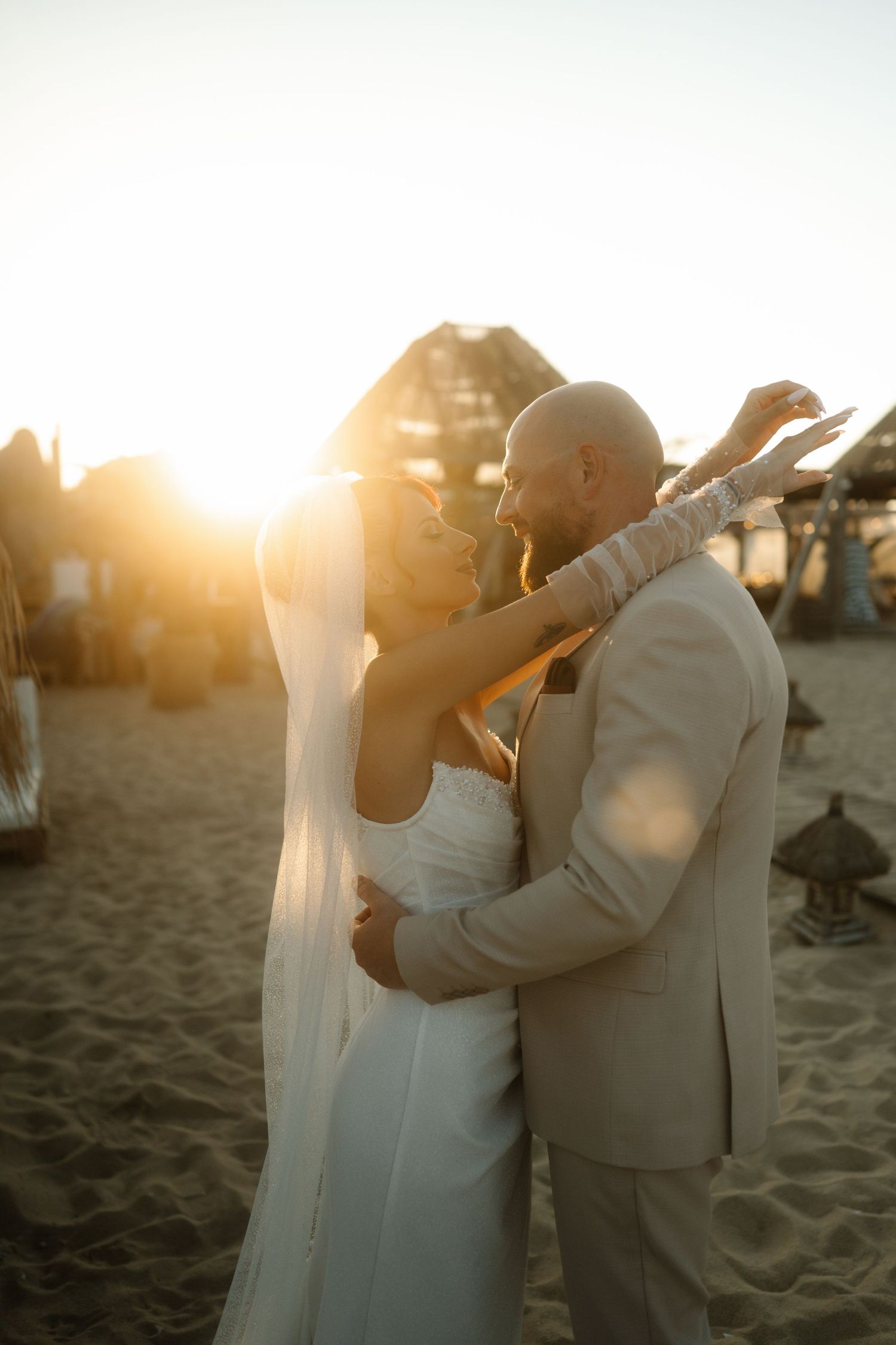 A romantic beach wedding couple embracing during golden hour sunset with warm glowing light behind them.