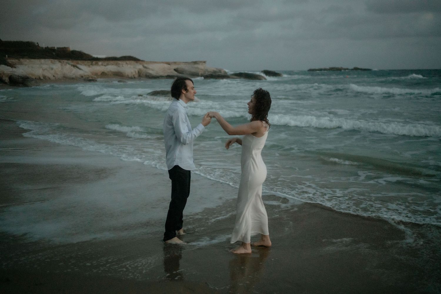 Couple dancing romantically on a beach shore with ocean waves and rocky cliffs in the background.