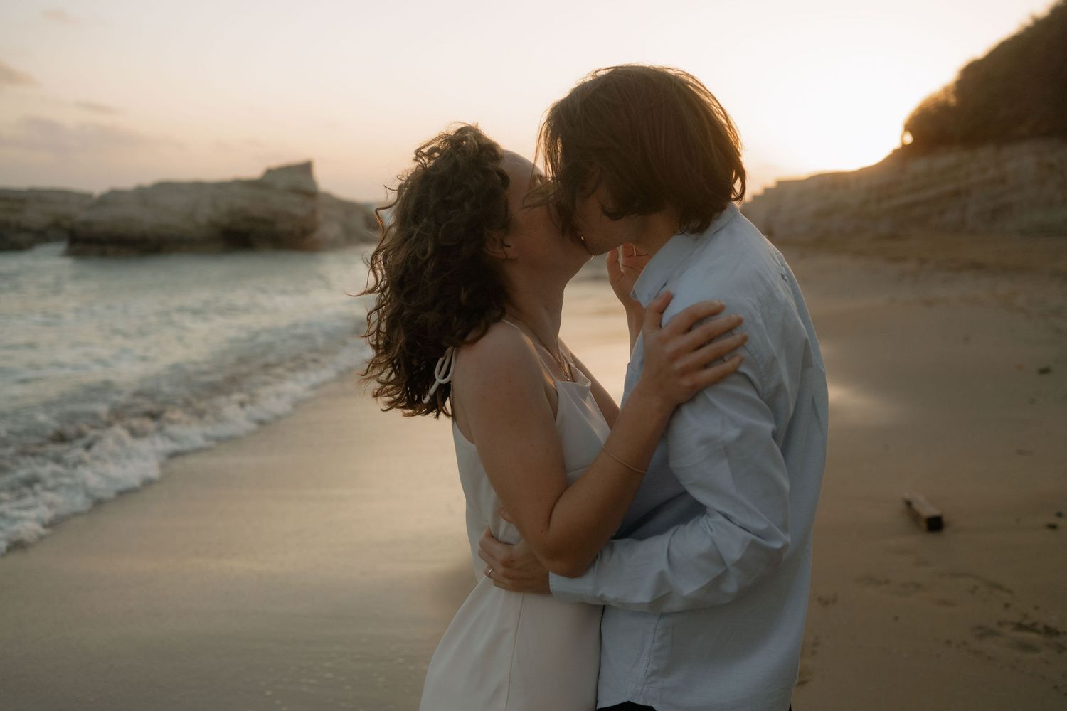 A couple shares a romantic kiss on a sandy beach at sunset with rocky cliffs in the background.