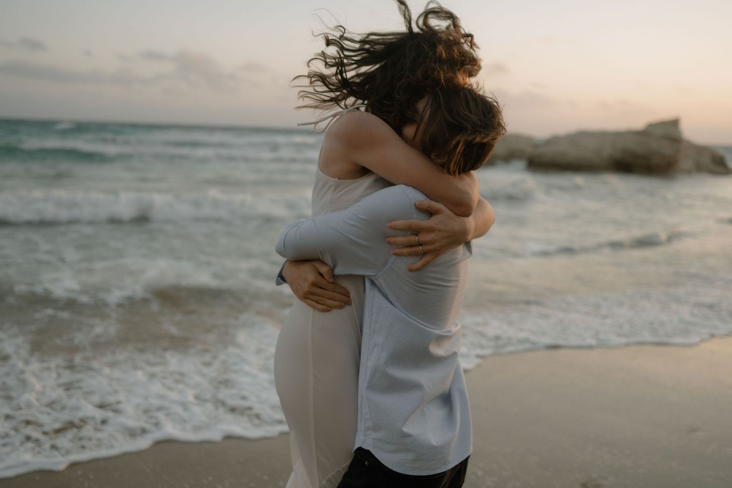 Couple embracing romantically on a sandy beach at sunset with ocean waves in the background.