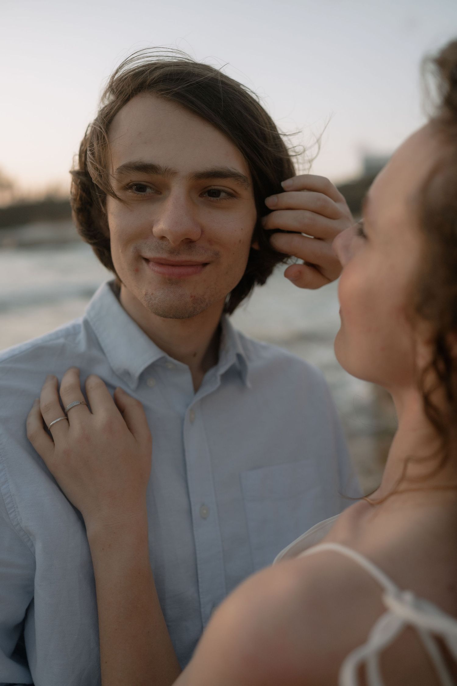 A couple shares a tender moment at sunset on a beach, with the woman touching the man’s hair as he smiles warmly.