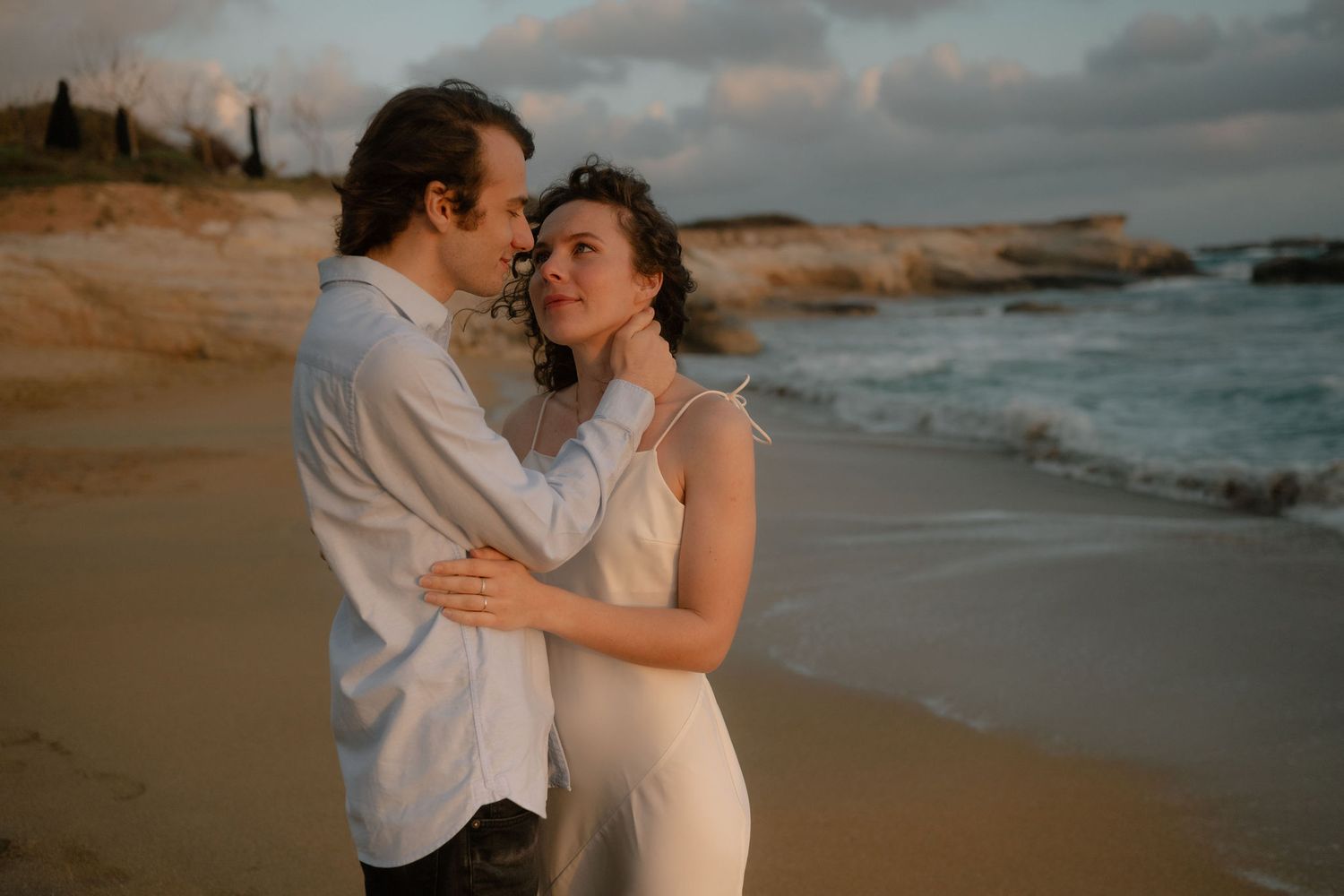 Couple embracing on a sandy beach at golden hour, waves crashing behind them, woman in white dress.