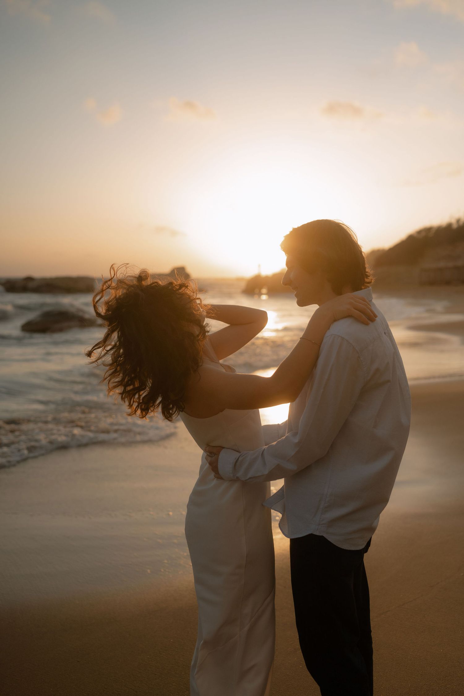 Couple embracing on a sandy beach at golden sunset, waves crashing behind them, woman's hair blowing in the wind.