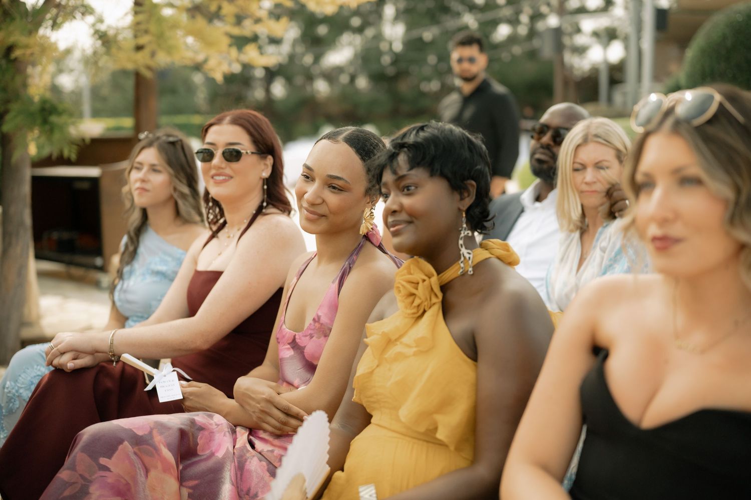 Diverse group of elegantly dressed women seated outdoors at a wedding ceremony, smiling and enjoying the celebration.