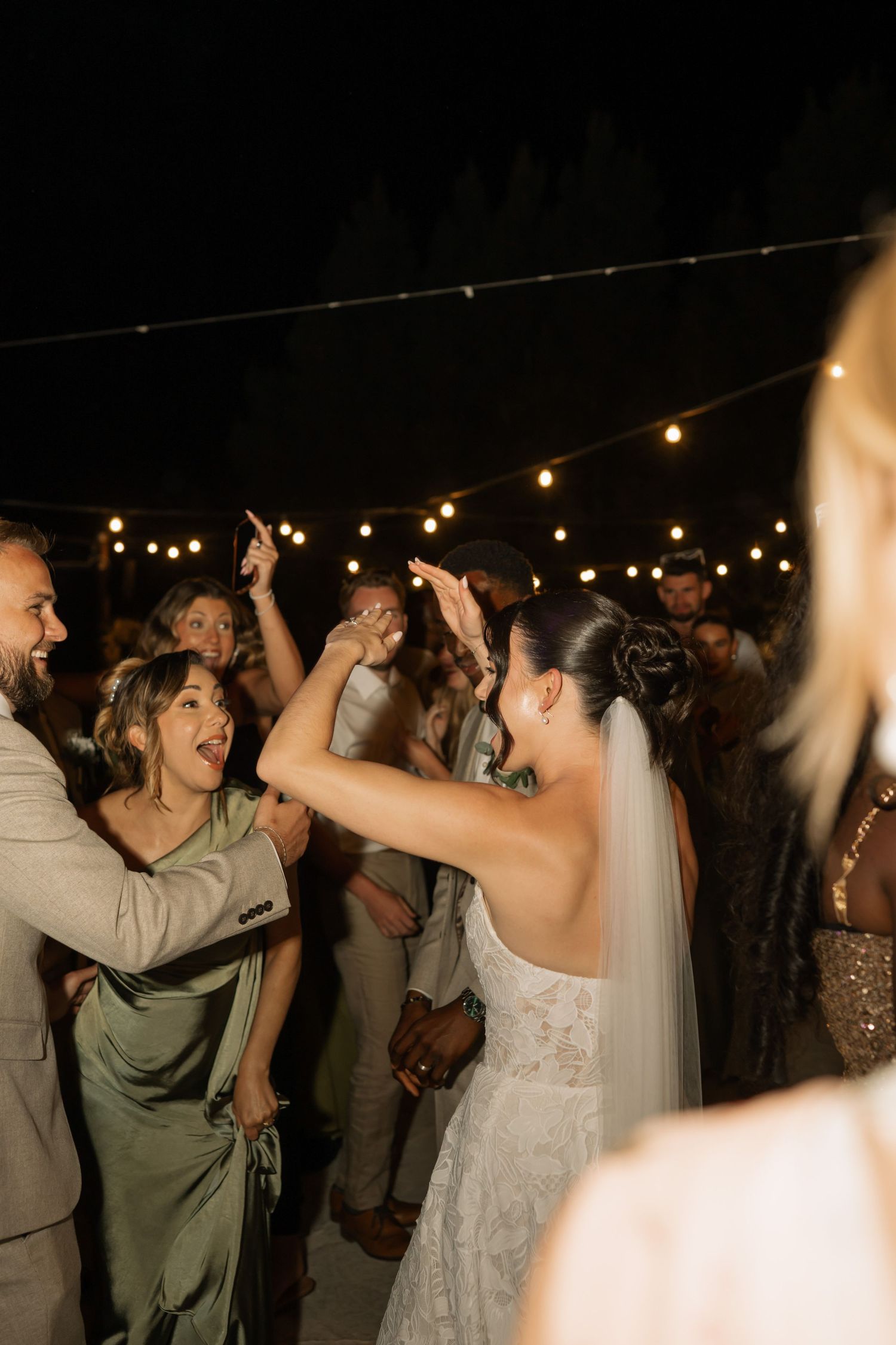 Bride in lace wedding dress and veil dancing with guests under string lights at an outdoor evening wedding reception.