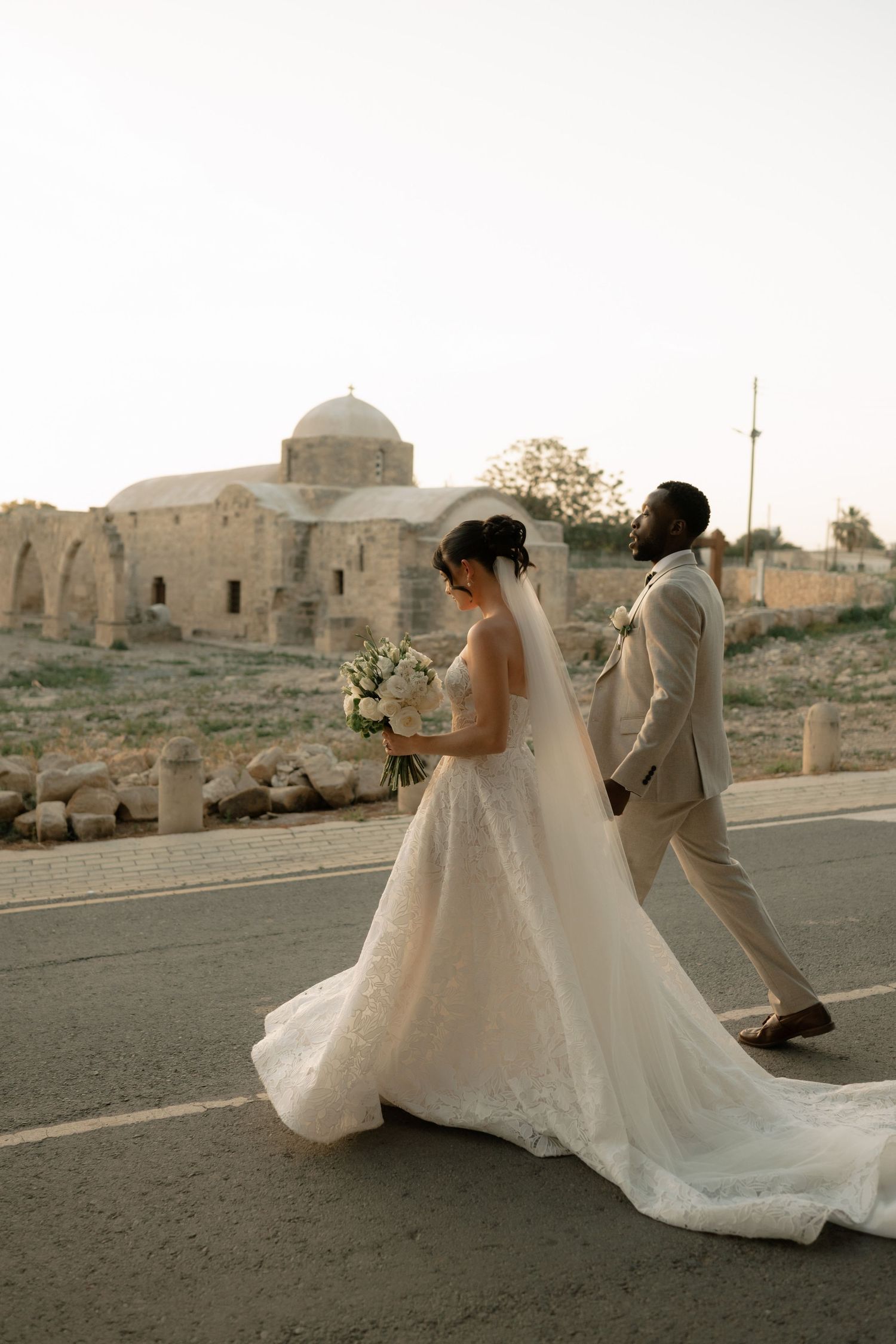 Bride and groom walking past ancient stone church ruins on their wedding day, bride in lace gown with veil and bouquet.