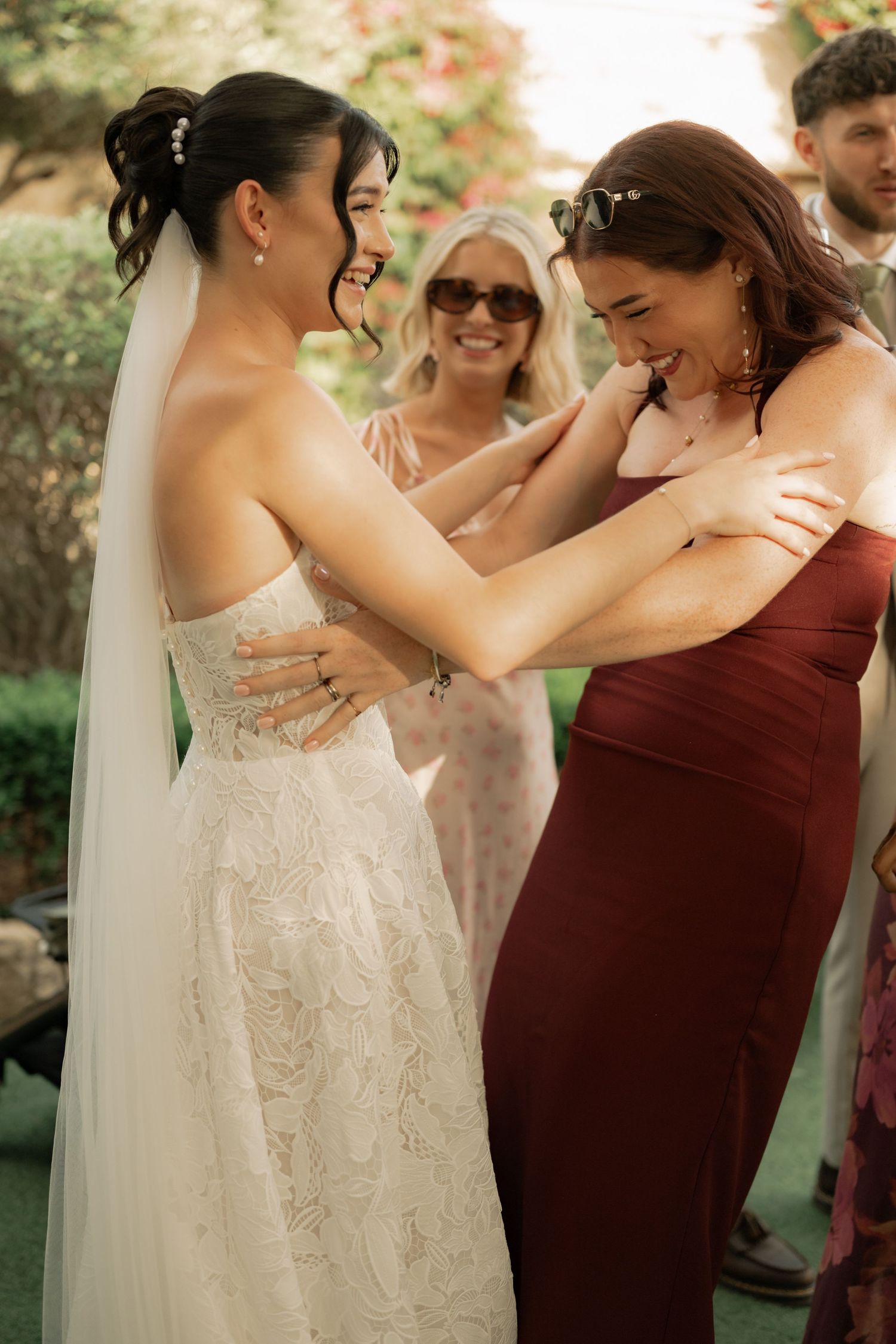 Bride in white lace gown and veil shares a joyful moment with bridesmaid in burgundy dress at outdoor wedding.