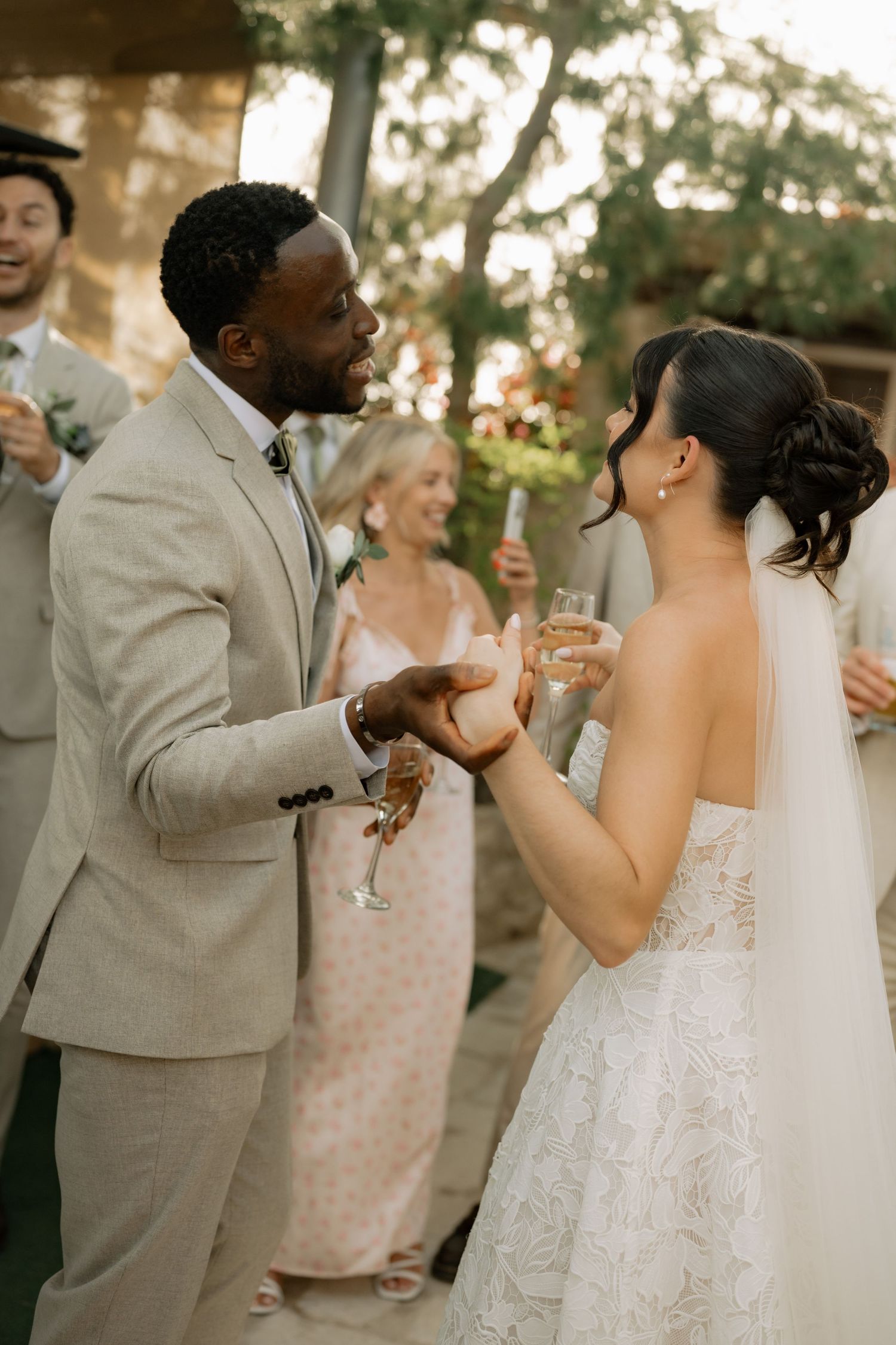 Bride and groom sharing a champagne toast at outdoor wedding reception, surrounded by smiling guests in elegant attire.