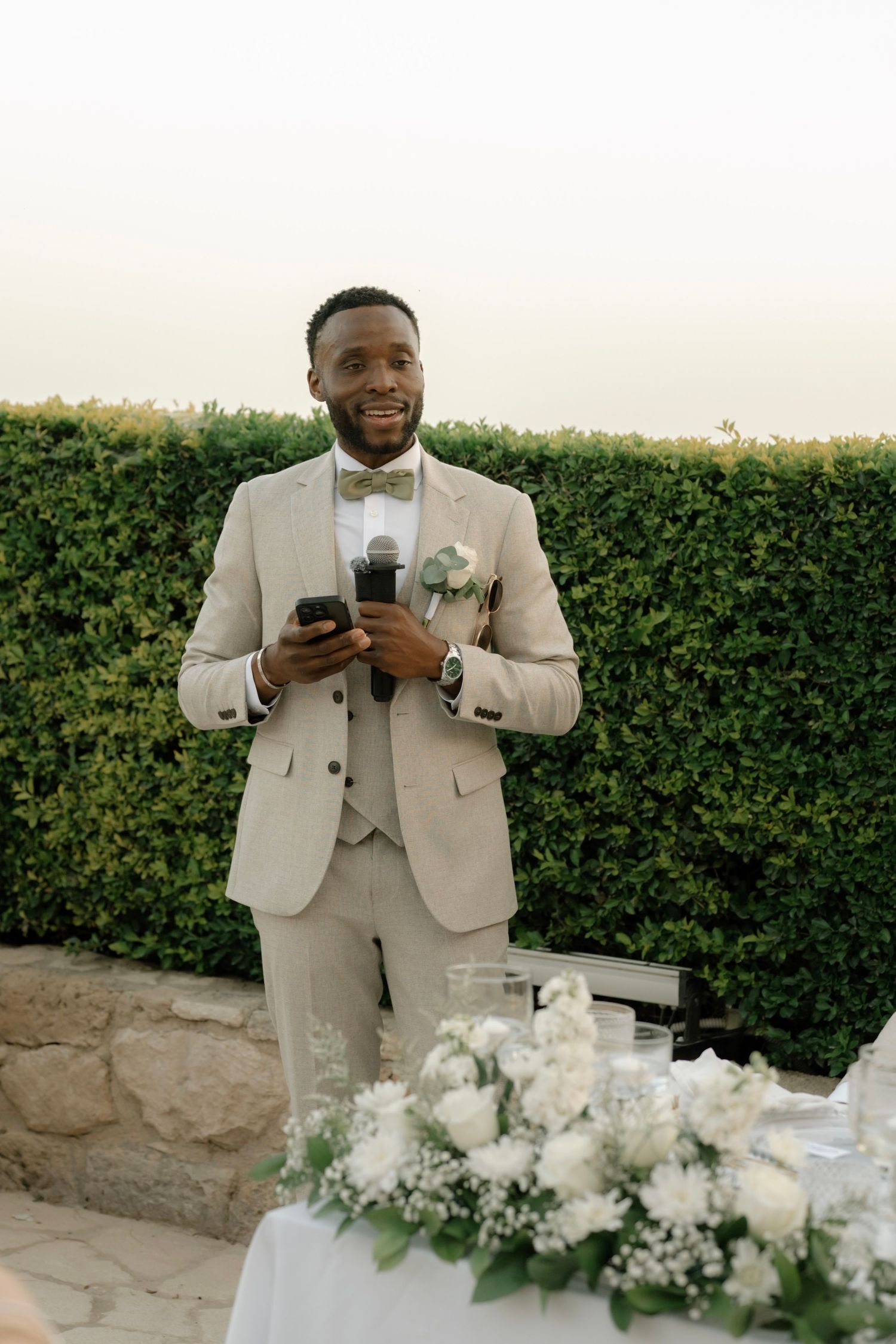 Smiling groom in beige suit holding microphone and phone, giving wedding speech outdoors near green hedge.