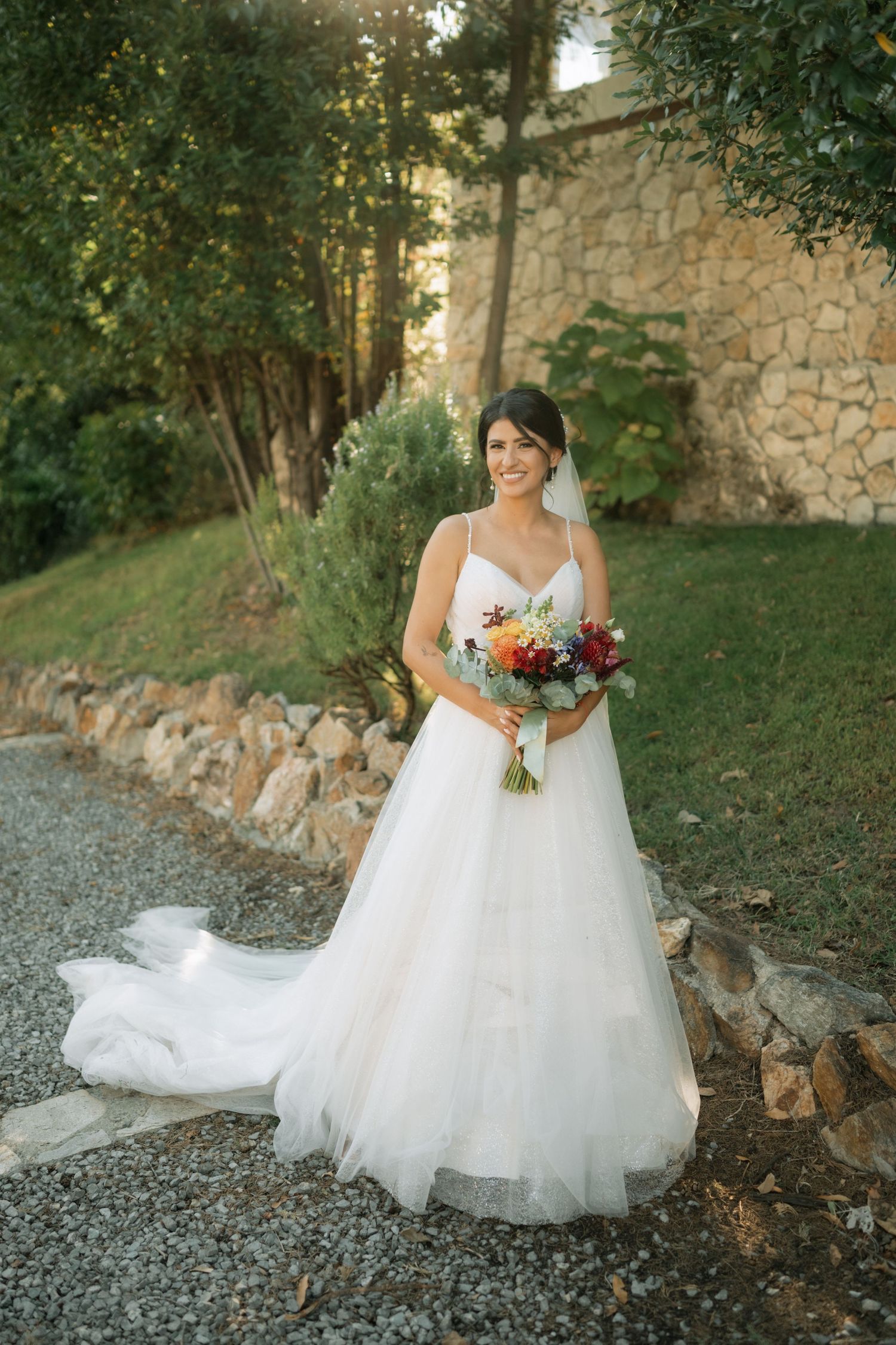 A beautiful white wedding dress flows across a rustic stone path surrounded by lush greenery and stone walls.