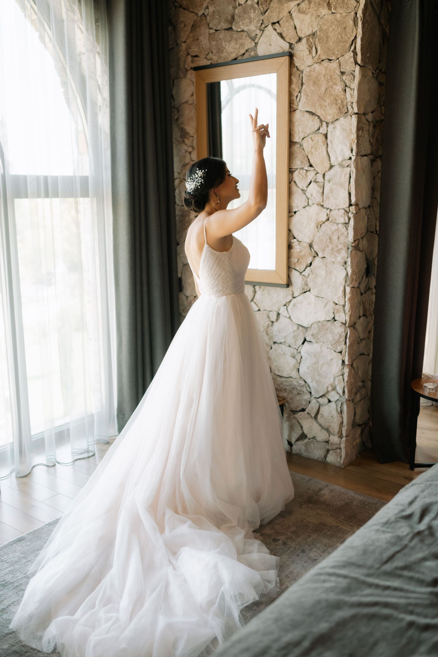 A bride in a flowing white wedding gown stands by a stone wall, reaching up to touch a framed artwork.