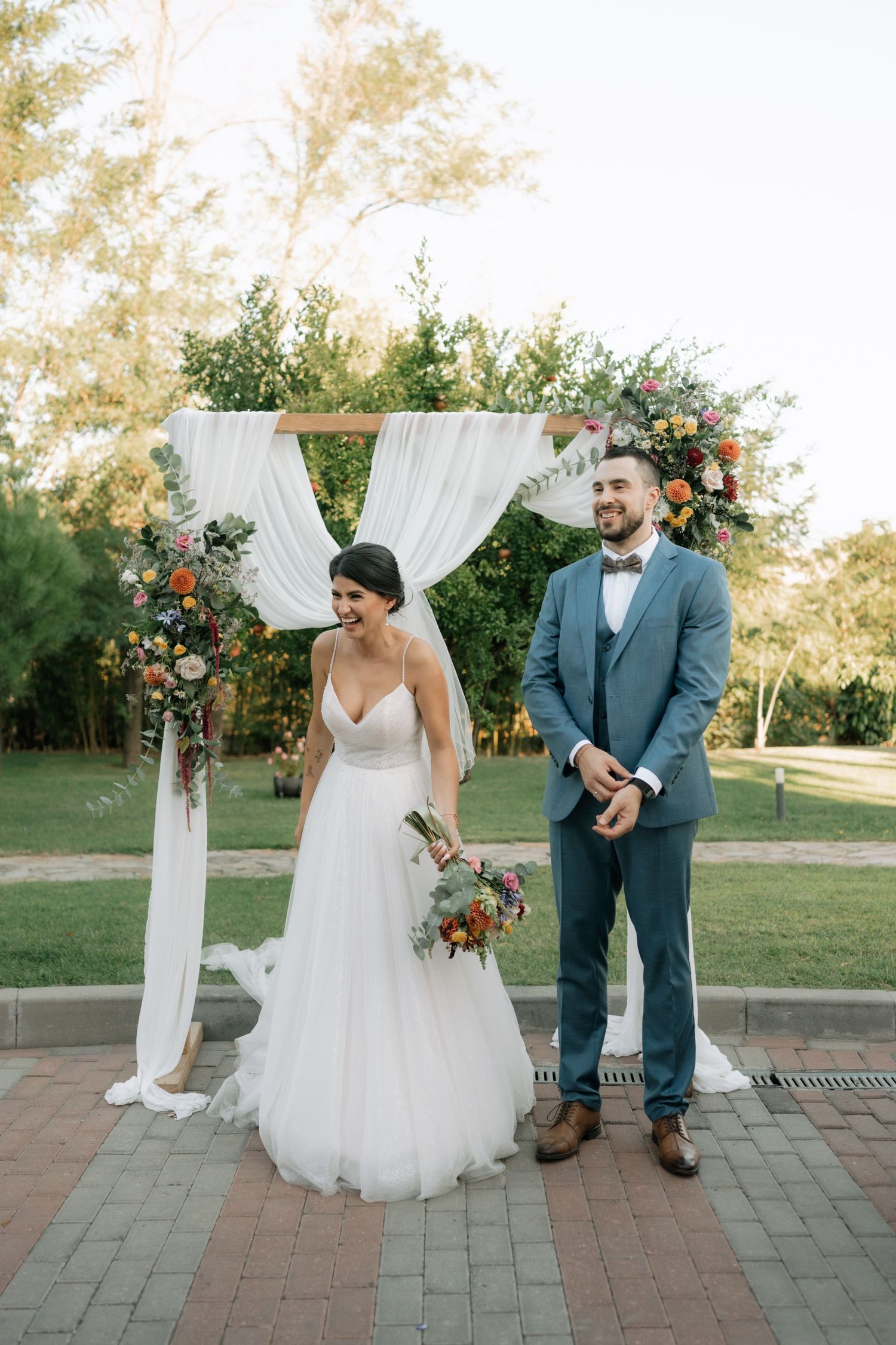 A romantic outdoor wedding ceremony on a brick patio with a white draped arch and floral decorations at sunset.