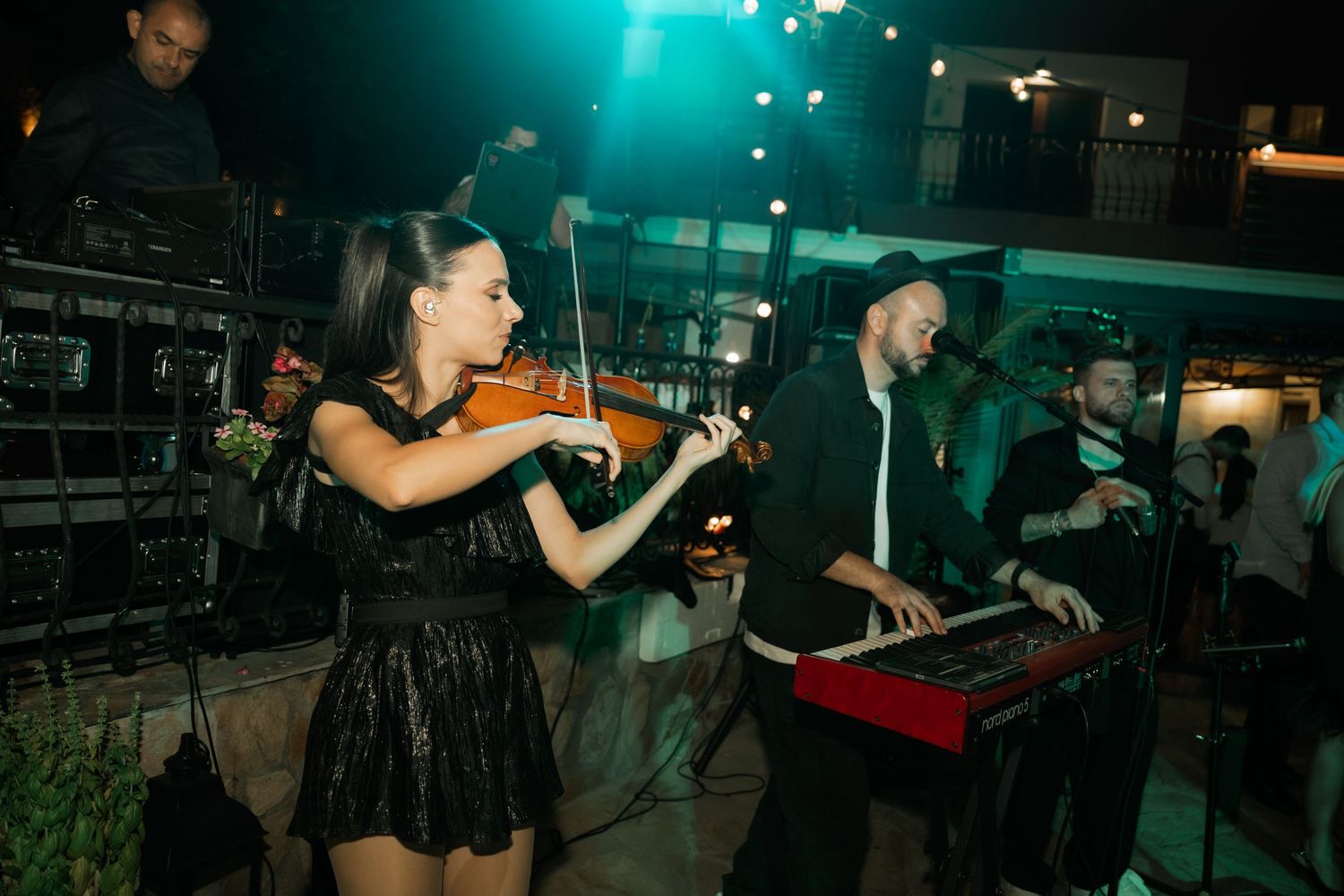 Musicians perform on stage at a dimly lit concert venue with teal lighting and glowing spotlights in the background.