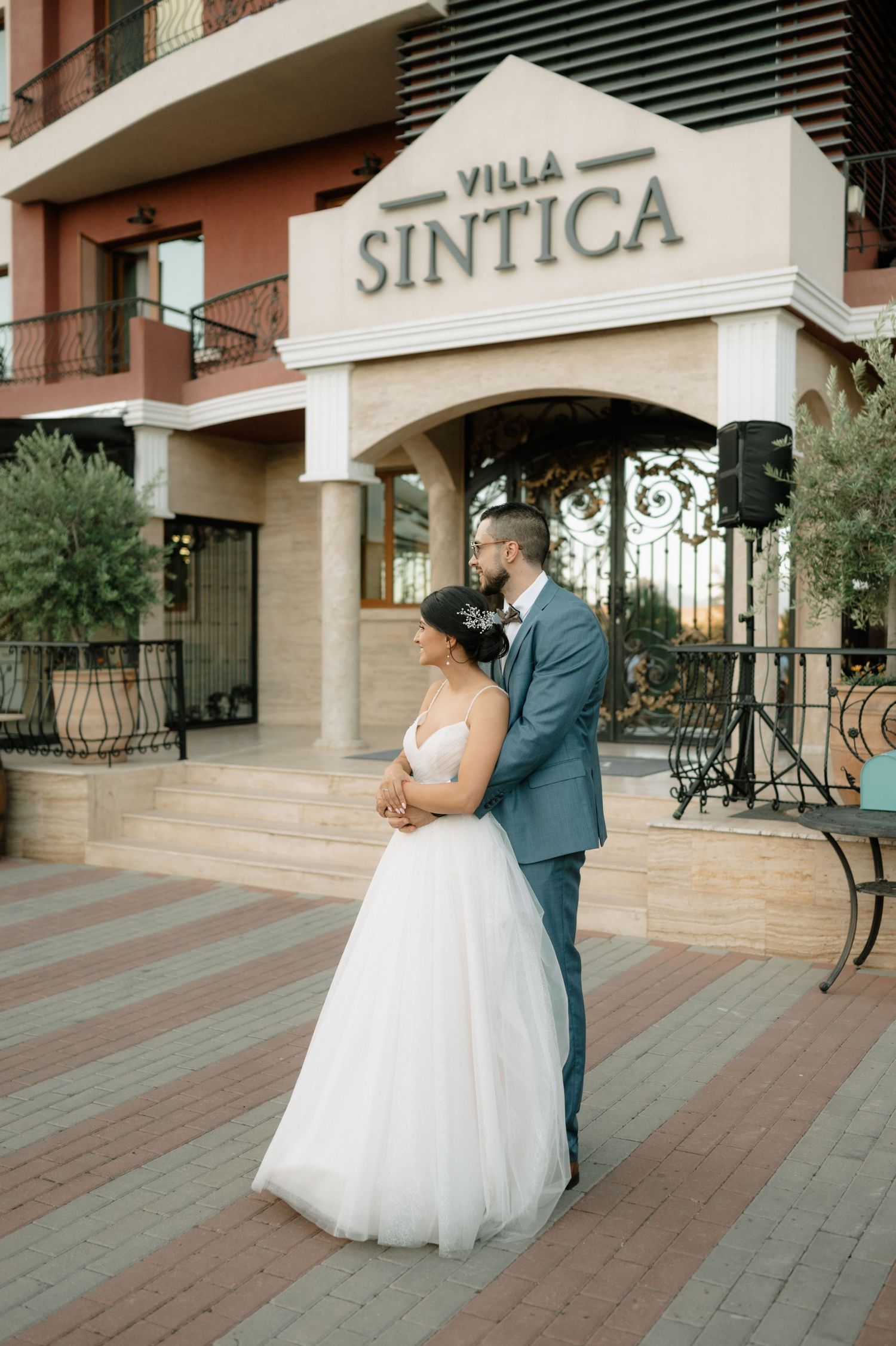A couple embraces outside Villa Sintica entrance with elegant wedding attire against a terracotta building facade.