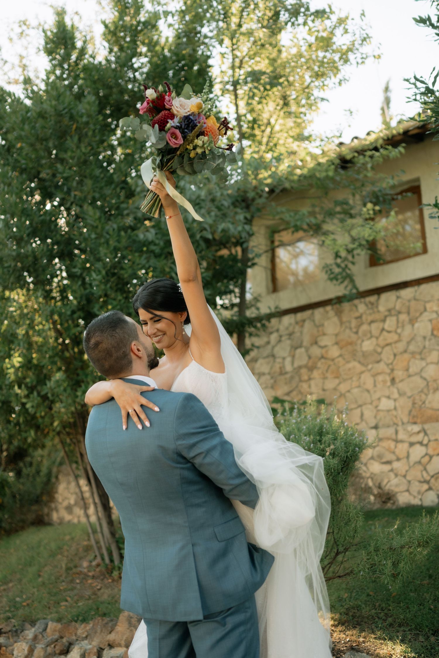 Newlyweds dance together in a romantic garden setting with stone wall backdrop and natural sunlight.