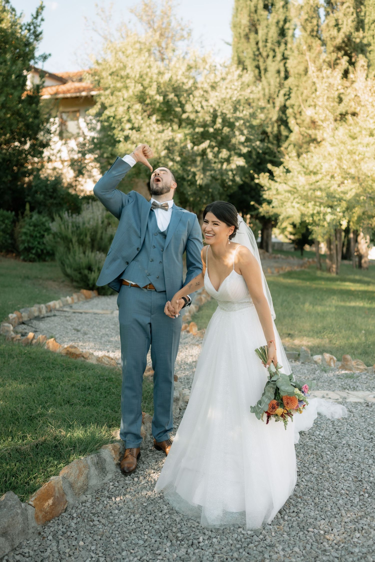 A couple in wedding attire share a playful moment walking together on a gravel path surrounded by autumn trees.