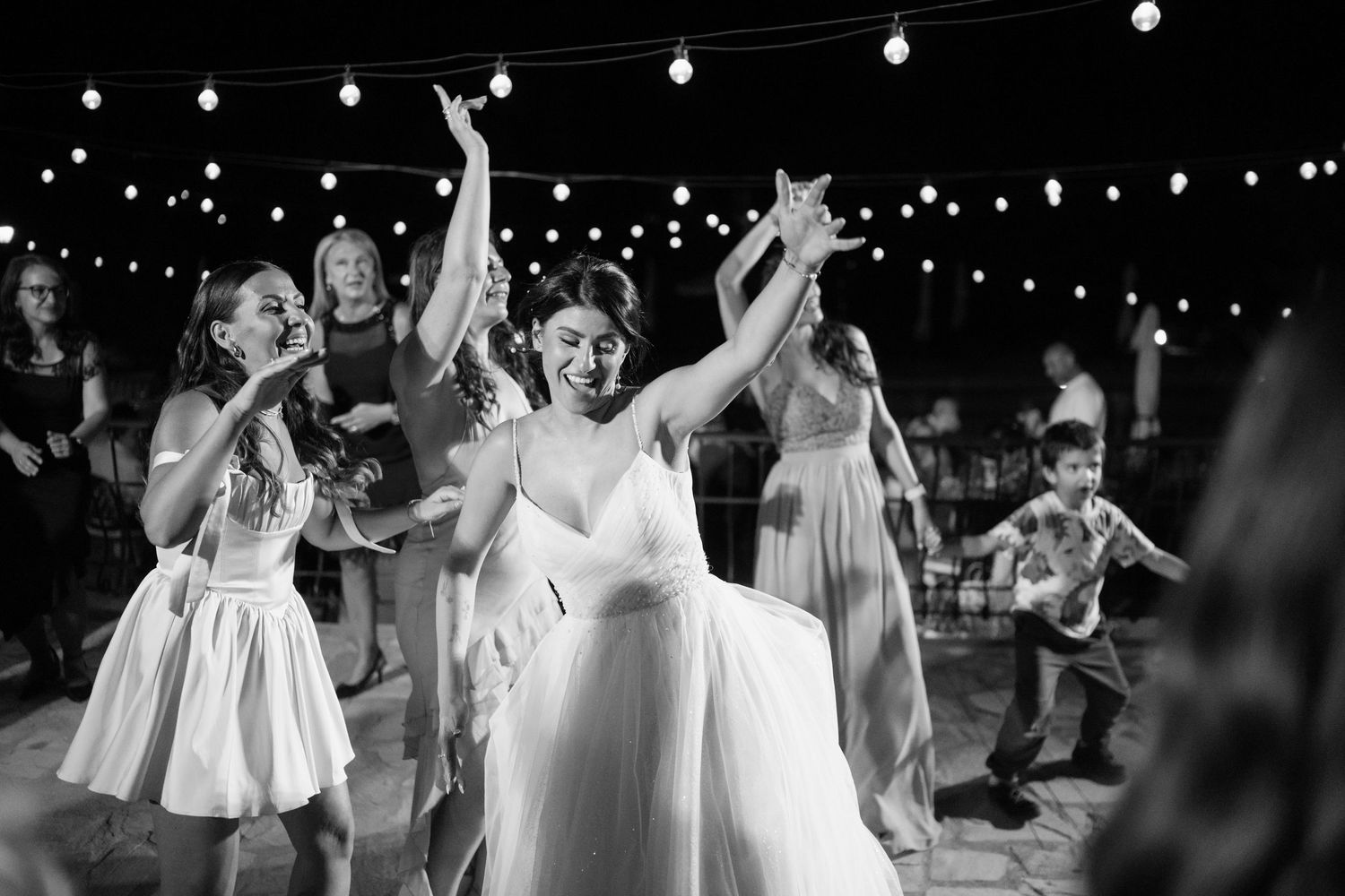 Wedding guests dancing and celebrating on an outdoor dance floor illuminated by overhead string lights at night.