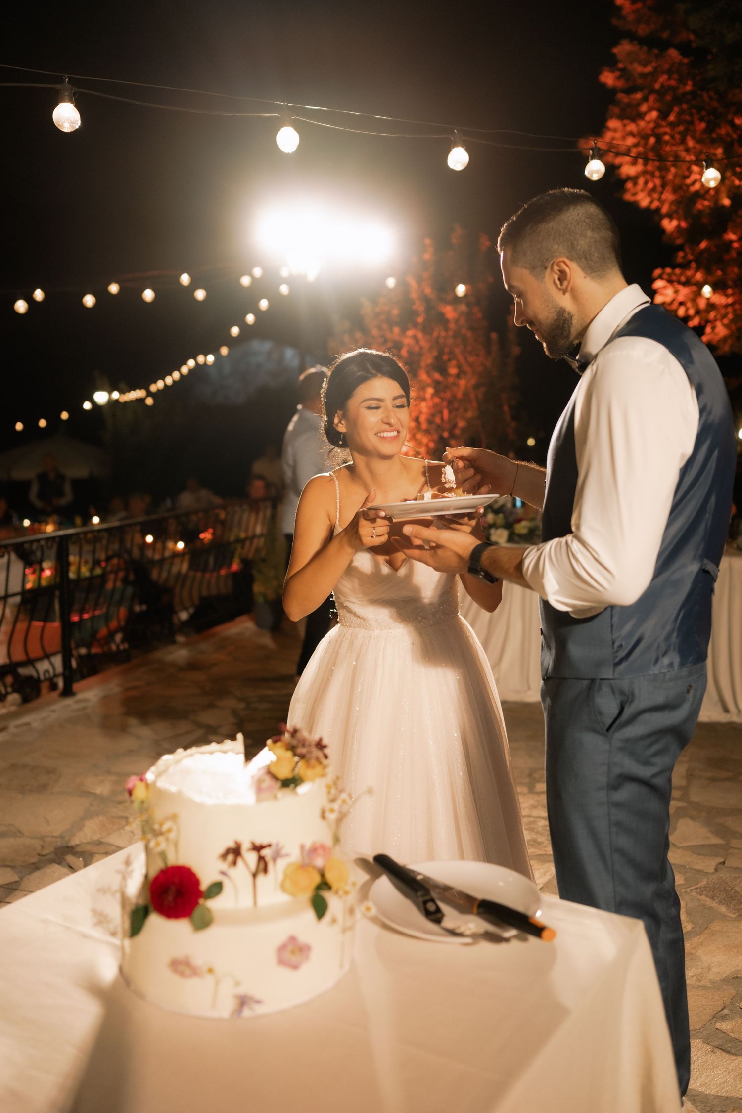 A couple shares a playful moment while cutting their wedding cake together under twinkling outdoor string lights at night.