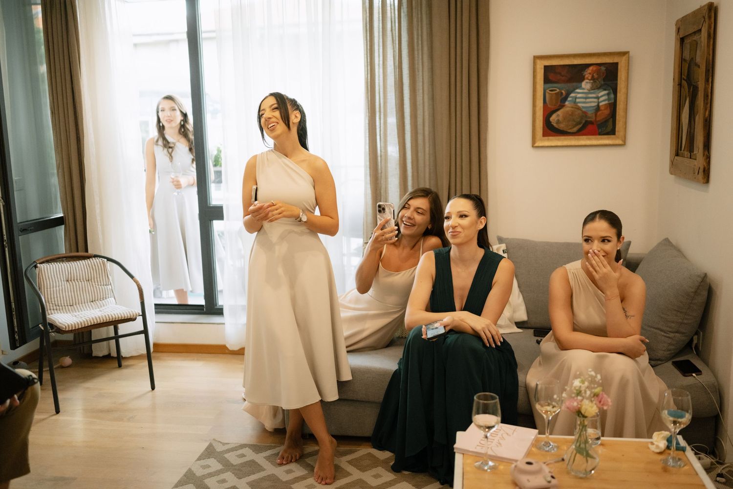 Bridesmaids in elegant dresses share emotional reactions while getting ready in a bright, modern hotel room.